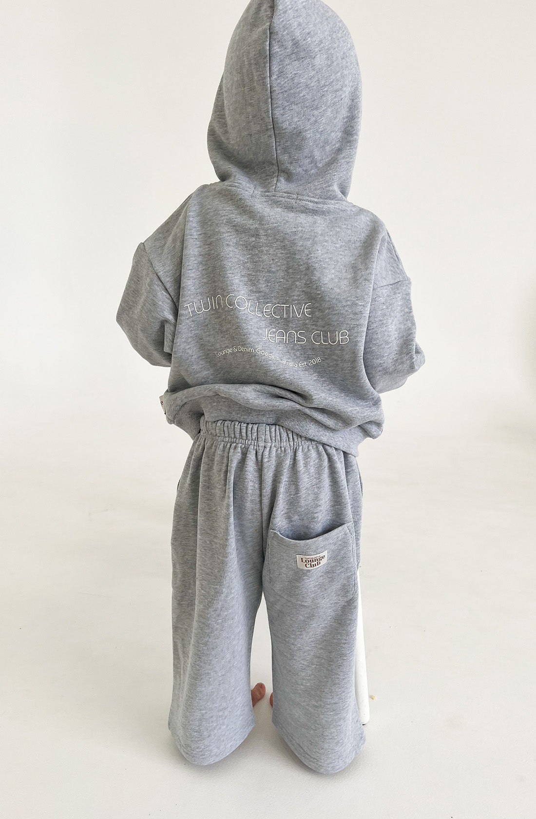 A child stands facing away, wearing a grey TWIN COLLECTIVE Logo Hoodie Set with the hood up and matching oversized track pants. The hoodie features text on the back that reads "Twin Collective Jeans Club." The child's attire is casual and comfortable, set against a plain white background.