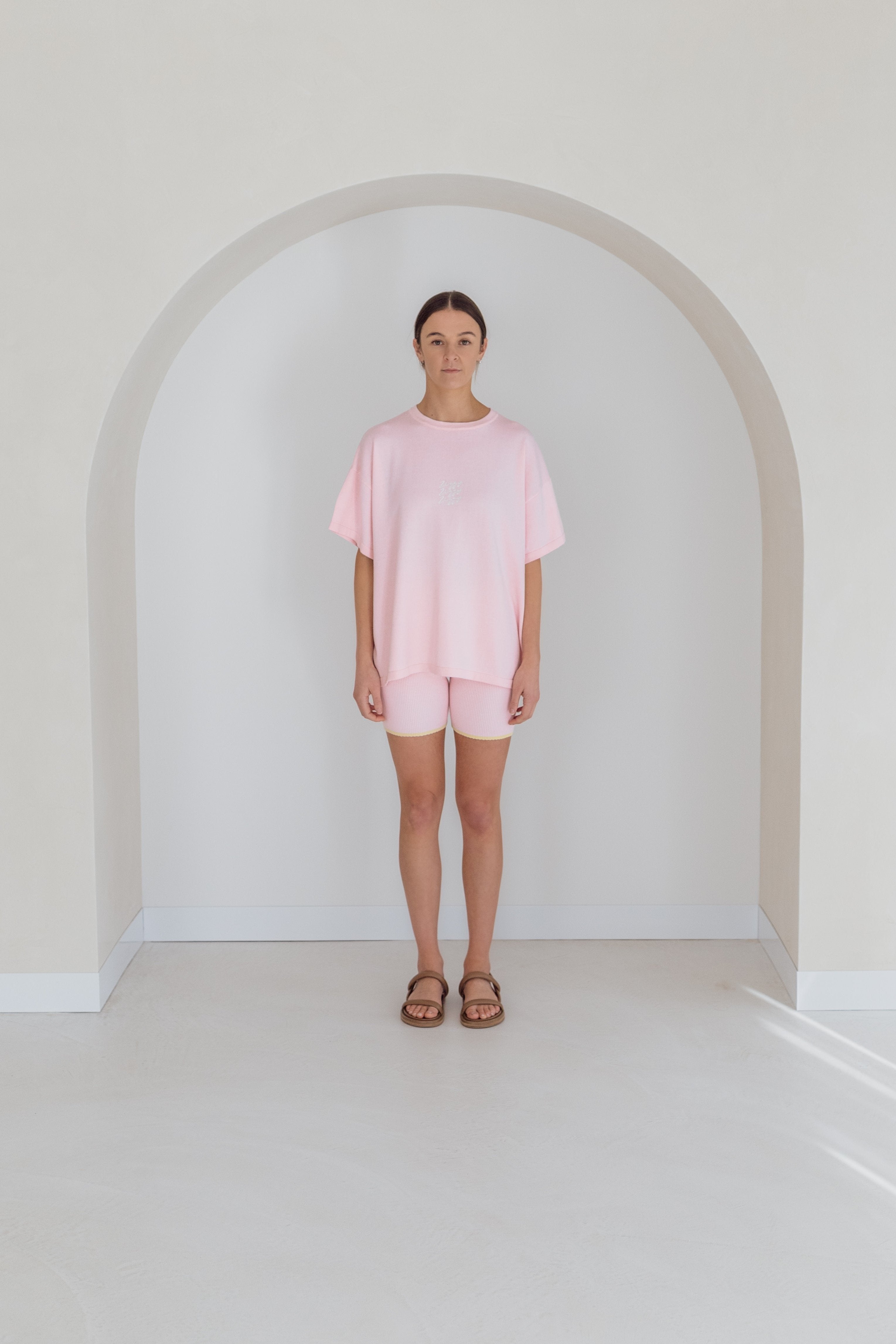 A person stands under a white archway wearing a pastel pink oversized Women's Signature Tee Freya by ZIGGY LOU, complete with matching shorts. They pair this outfit with brown sandals on a light-toned floor, complemented by a neutral wall in the background.