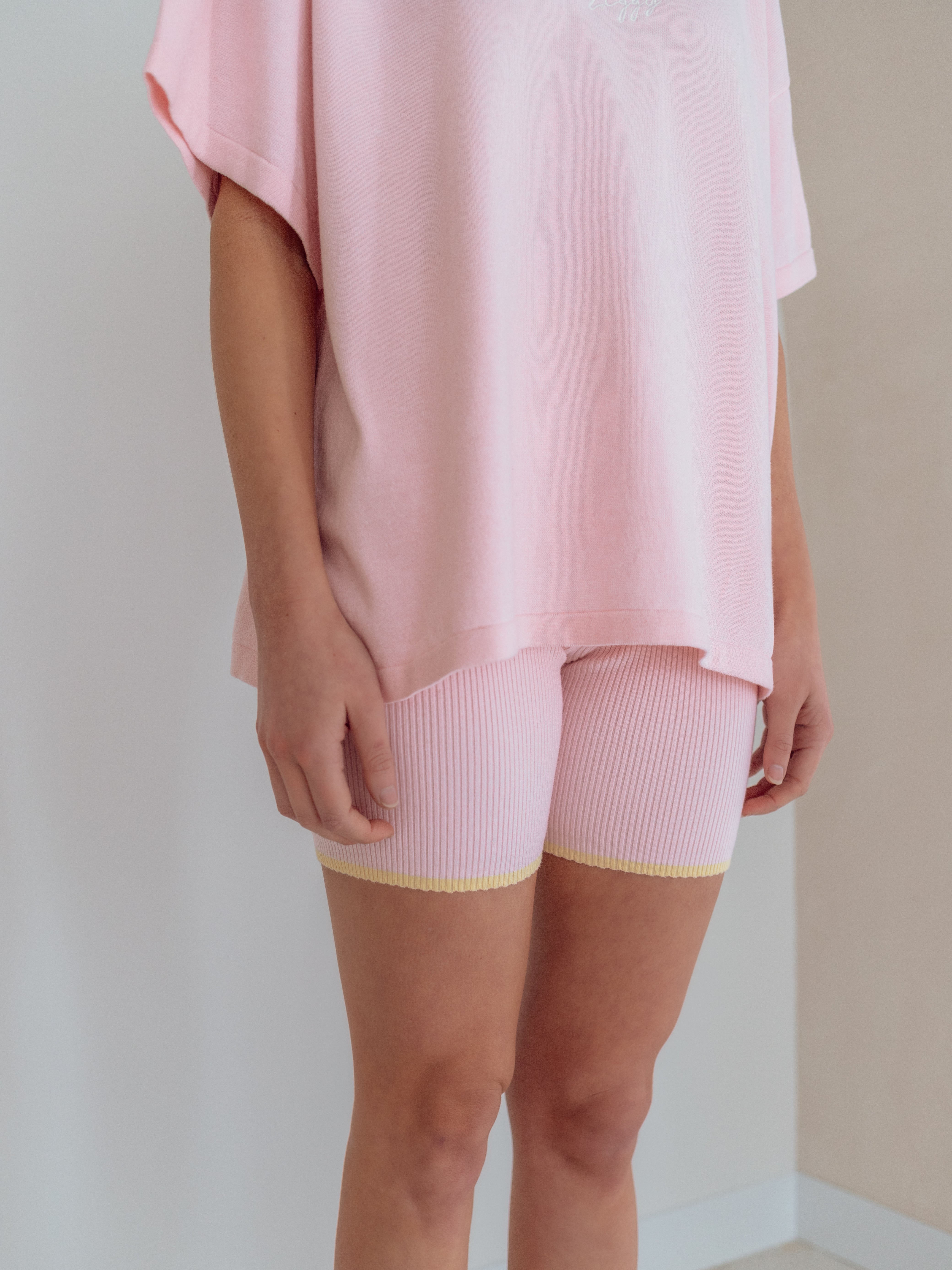 A person is wearing an oversized pink Women's Signature Tee Freya by ZIGGY LOU, paired with light pink ribbed shorts featuring yellow trim. They are standing indoors against a neutral wall, with their arms relaxed at their sides. The outfit is the focal point.