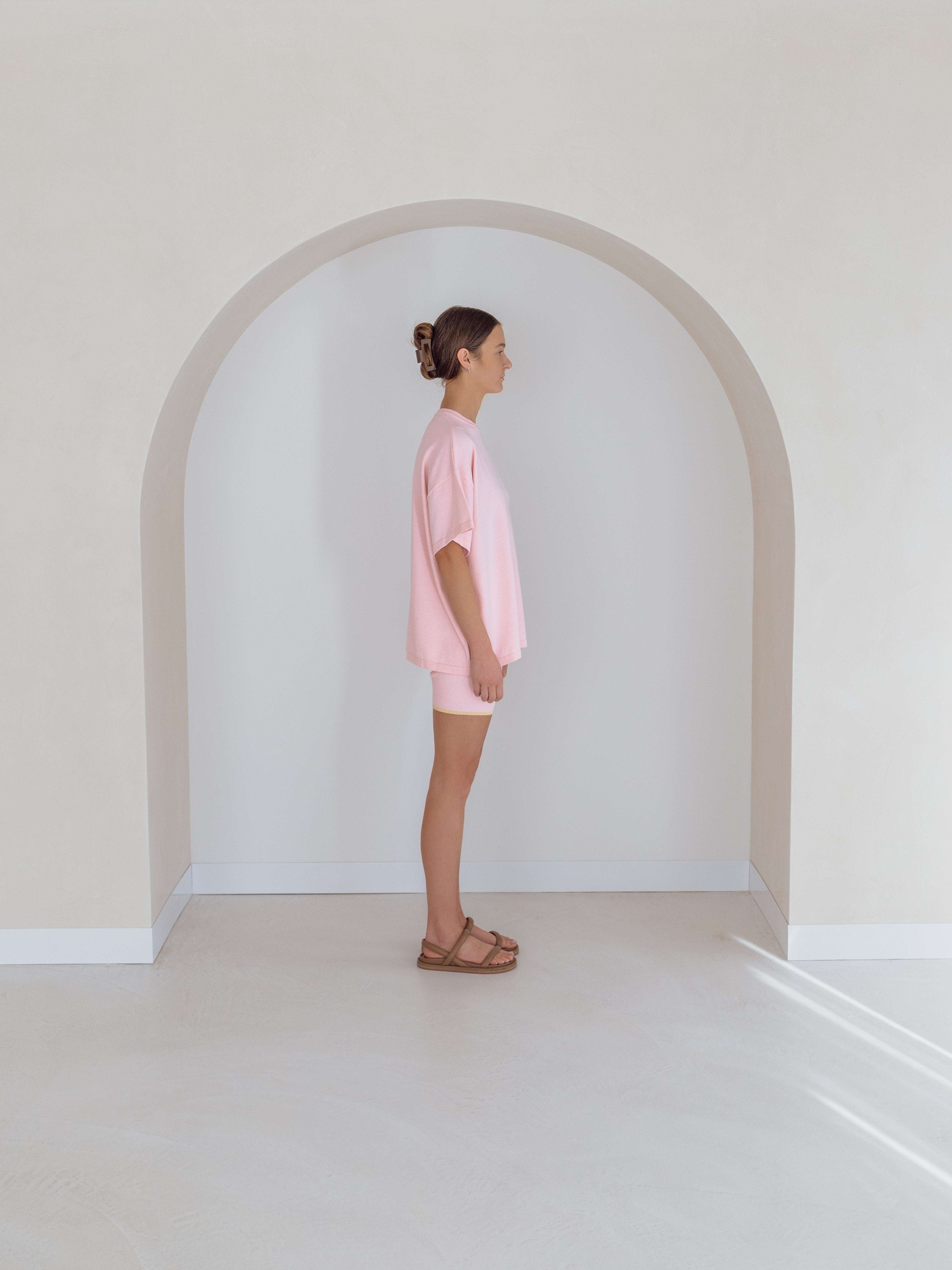 A person is standing sideways in a beige arched alcove, wearing the Women's Signature Tee Freya by ZIGGY LOU, which has an oversized fit in pink with the minimalist Ziggy embroidered logo. They are paired with shorts and brown sandals on a light floor.