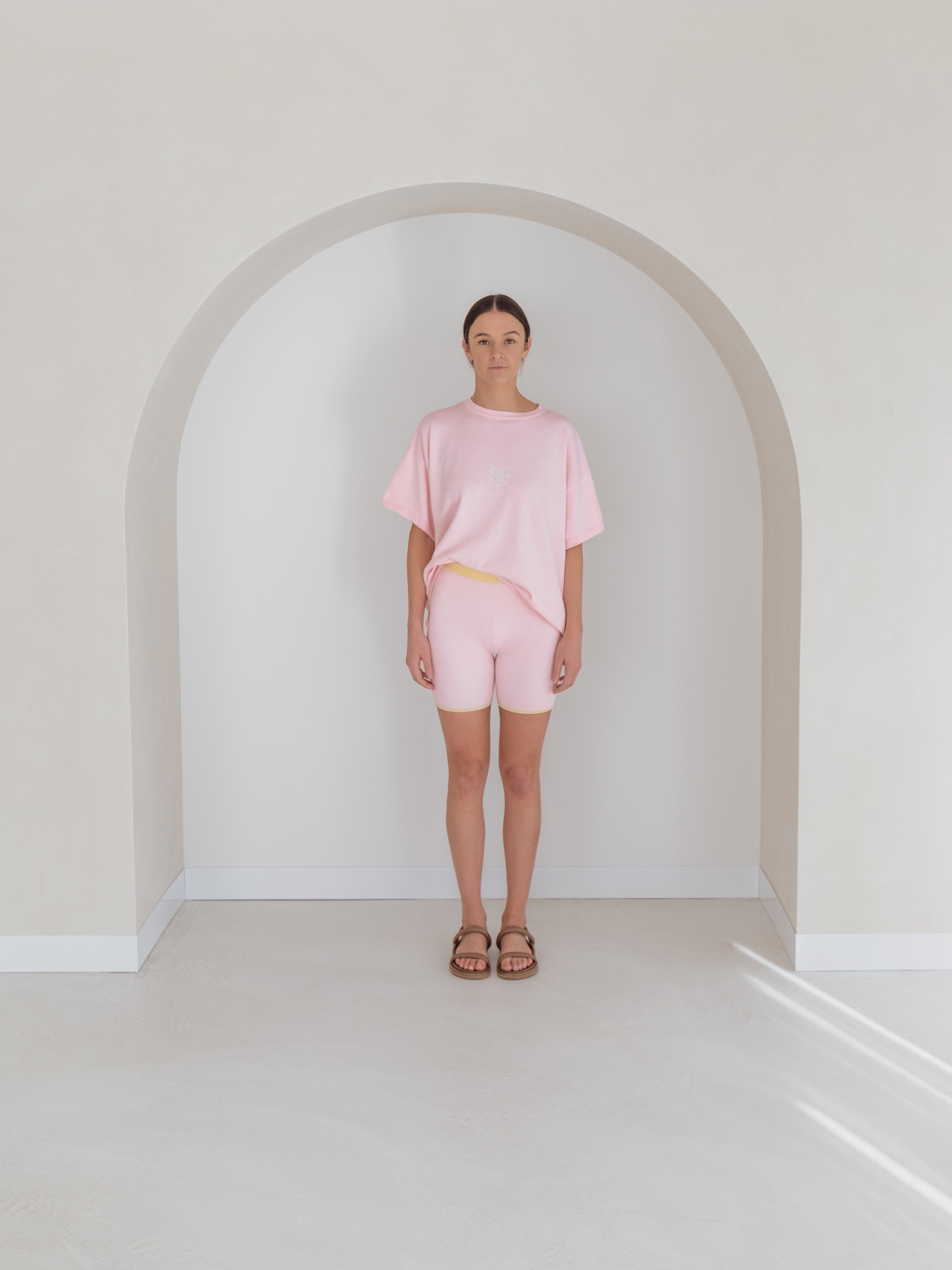 In a minimalist setting with a light-colored arched wall, an individual is dressed in an ethically crafted pink T-shirt paired with ZIGGY LOU's Women's Bike Shorts Clementine and brown sandals, presenting a relaxed and casual style. The overall ambiance is soft and neutral.