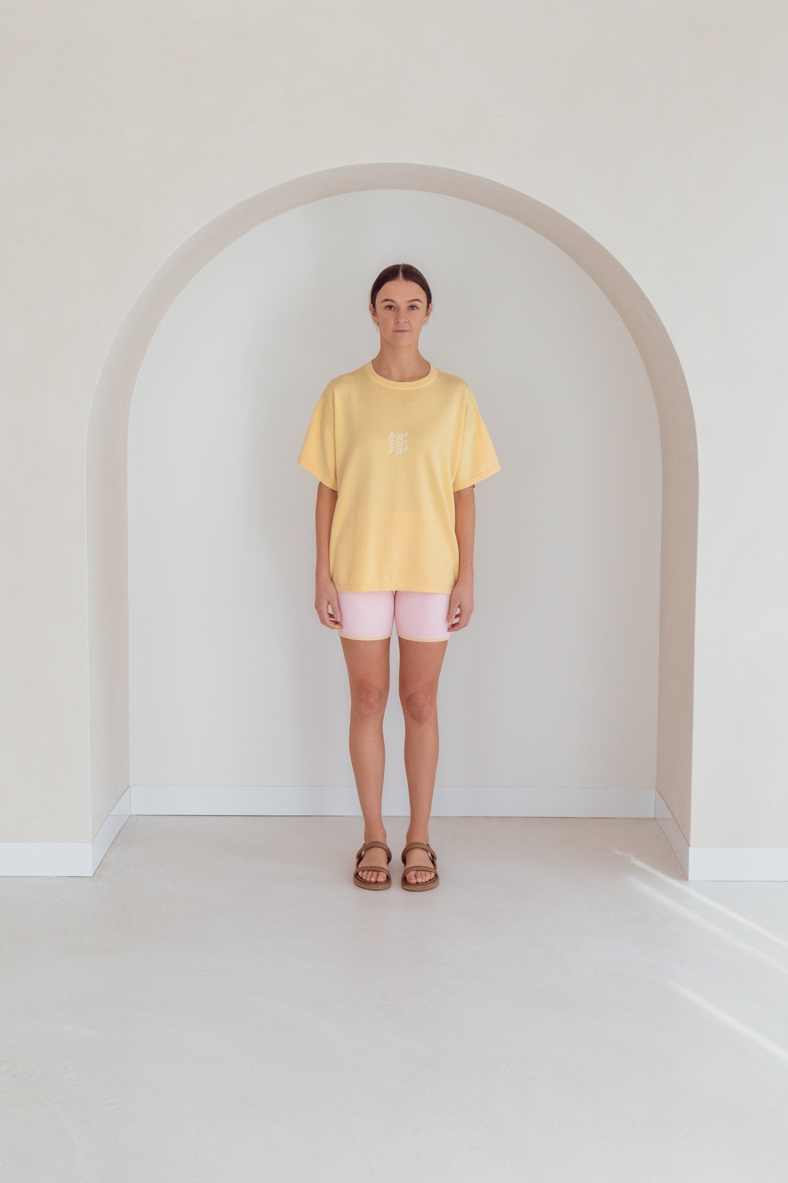 A person stands indoors under a white archway, wearing the oversized Women's Signature Tee Butter from ZIGGY LOU, along with pink shorts and brown sandals, looking directly at the camera. The setting features a minimalistic design with soft, neutral tones.