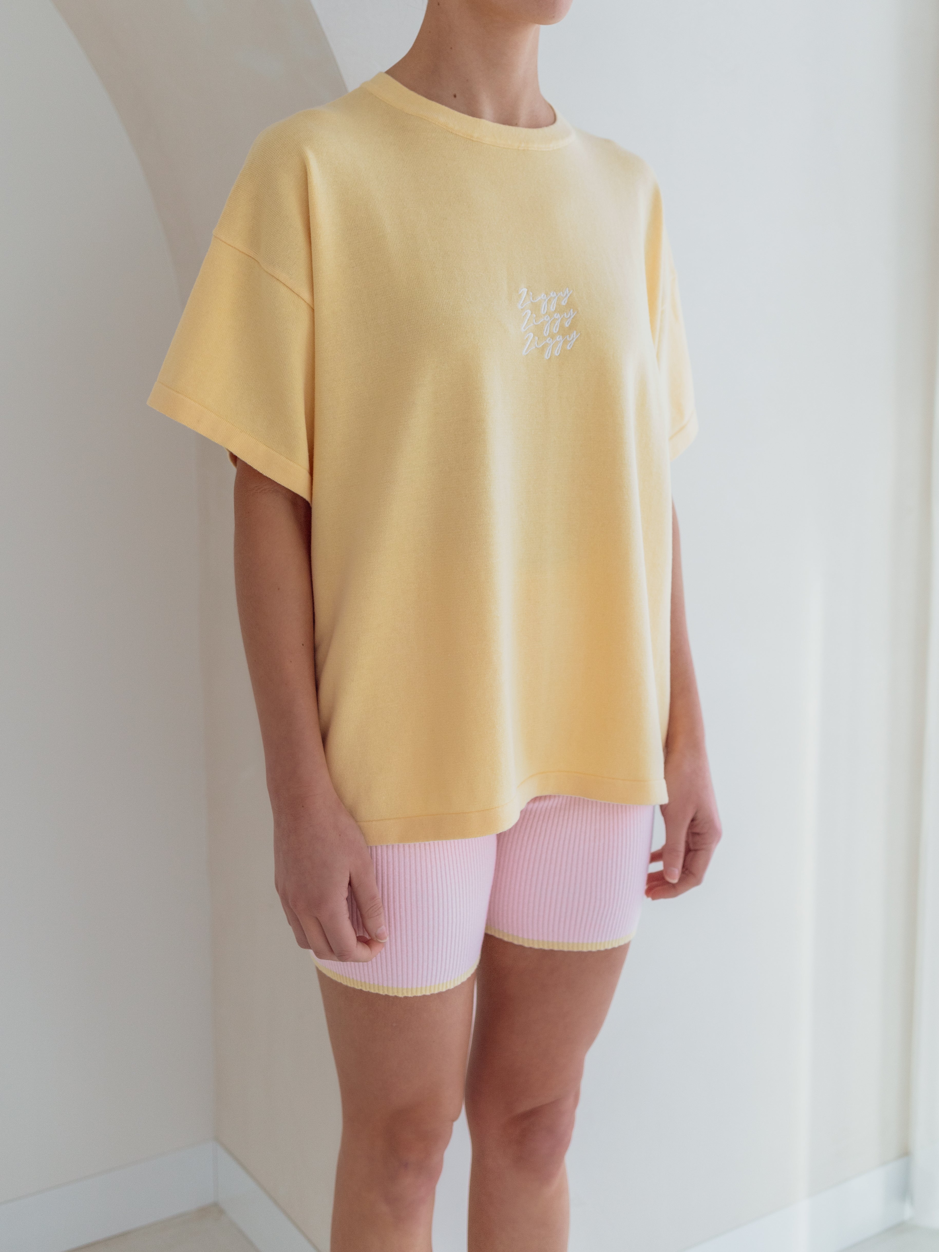 A person wearing an oversized Women's Signature Tee in Butter from ZIGGY LOU, paired with pink ribbed shorts, stands in front of a light-colored wall. The relaxed ensemble, softly lit by natural light, creates a casual yet stylish indoor scene.