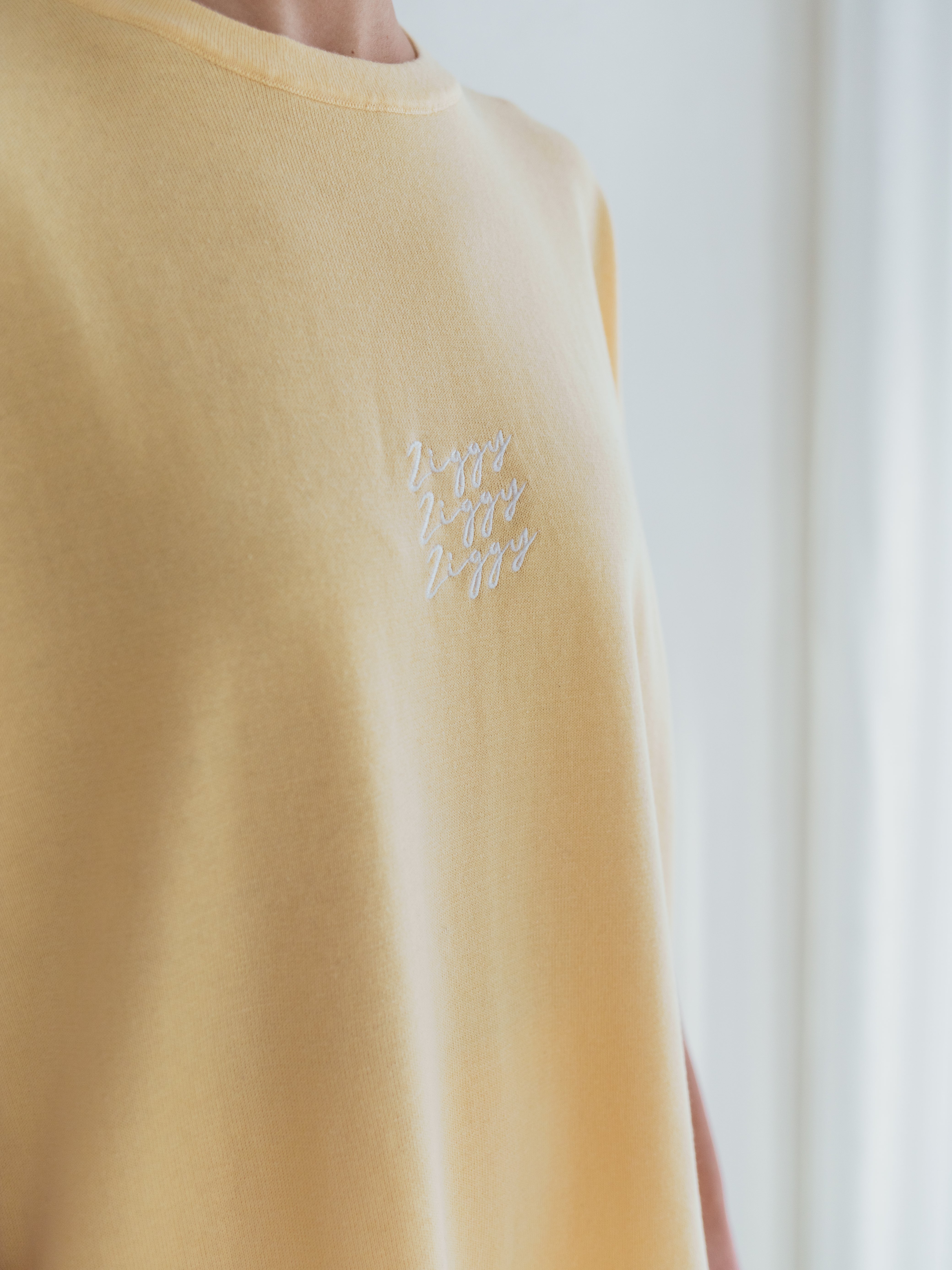 A person is wearing the Women's Signature Tee Butter from ZIGGY LOU, which is an oversized light yellow knitted tee featuring "Ziggy" embroidered multiple times in white on the chest. The softly blurred background suggests a bright and airy room, accentuating the comfort and style of this signature piece.