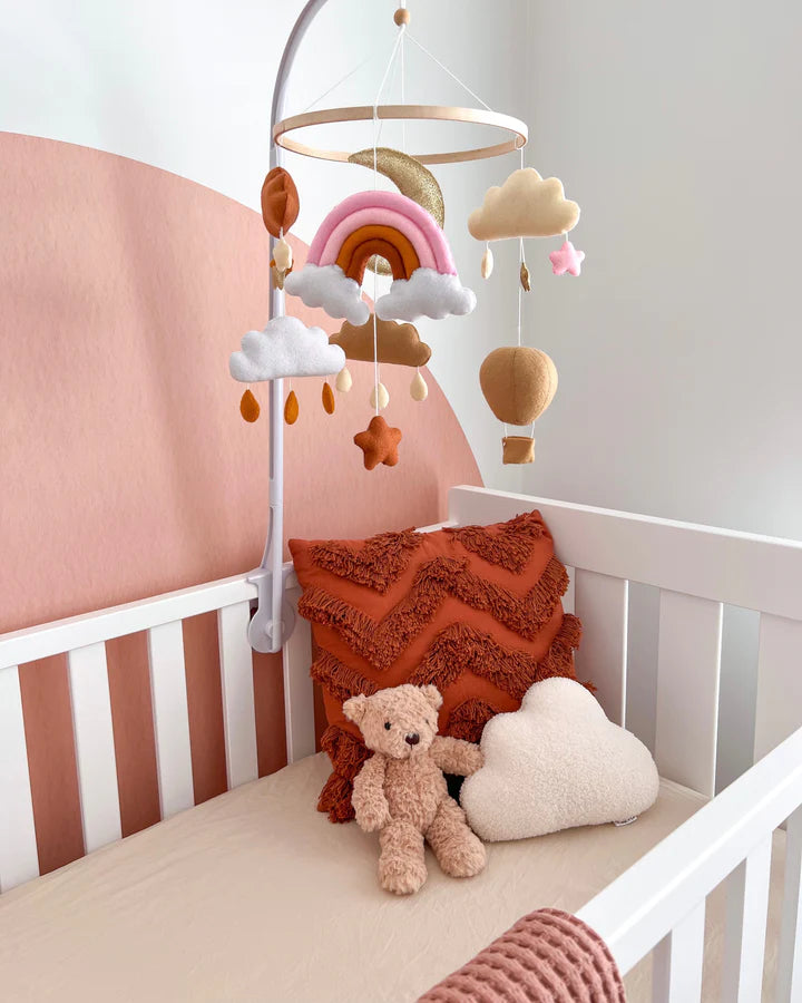 A white crib with rust zigzag pillows, a plush teddy bear, and a cloud cushion offers cozy comfort. Above, the ROOMI & ME Baby Mobile Sparkly Galaxy adds visual stimulation with felt clouds, rainbows, and hot air balloons against the pink accent wall.