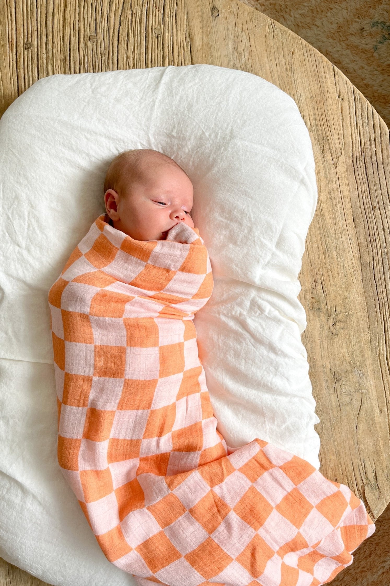 A baby is peacefully sleeping, wrapped in the Apricot Peaches Wrap with unique textile prints from MILKY DESIGNS. The baby is resting on a soft, white cushion placed on a wooden surface. The serene setting exudes warmth and comfort.