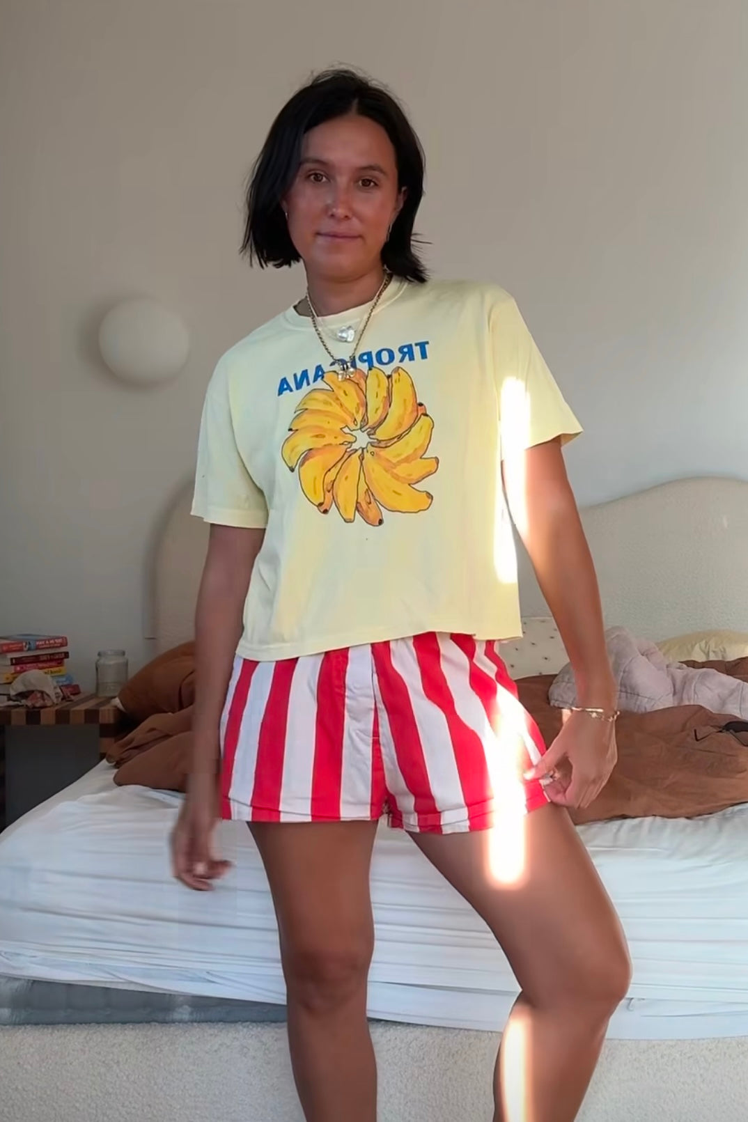 A person stands in a bedroom, wearing the Tropicana Banana T-Shirt by MAKU THE LABEL, a unisex tee made of 100% cotton paired with red striped shorts. The cozy room features a warm bed, a nightstand filled with items, and soft lighting.