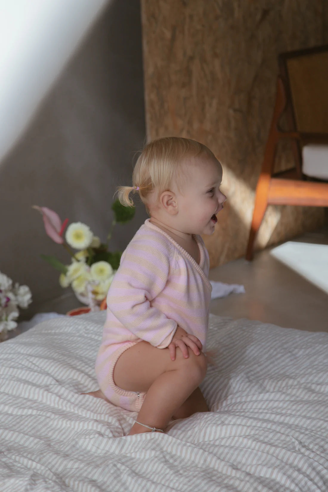 A baby wearing the Essential Wrap Romper Pink/Peach from MILKY DESIGNS smiles while kneeling on a bed. The background features a flower arrangement and a wooden chair. Warm light filters through, casting soft shadows, adding to the sense of comfort in the scene.