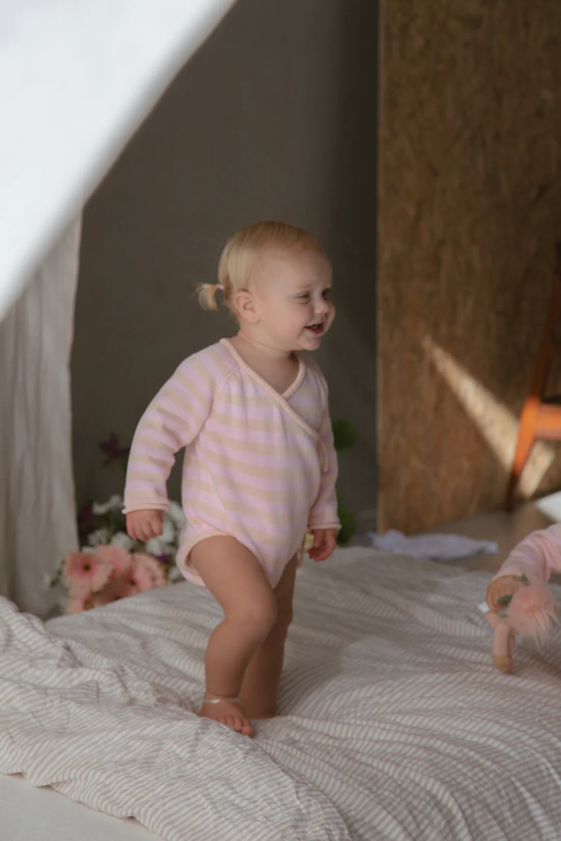 A blonde toddler stands on a beige and white striped bed, dressed in MILKY DESIGNS' Essential Wrap Romper Pink/Peach made from 100% cotton. The child sports a small ponytail and smiles while looking to the side. In the background, there are some flowers and a wall with wooden paneling.