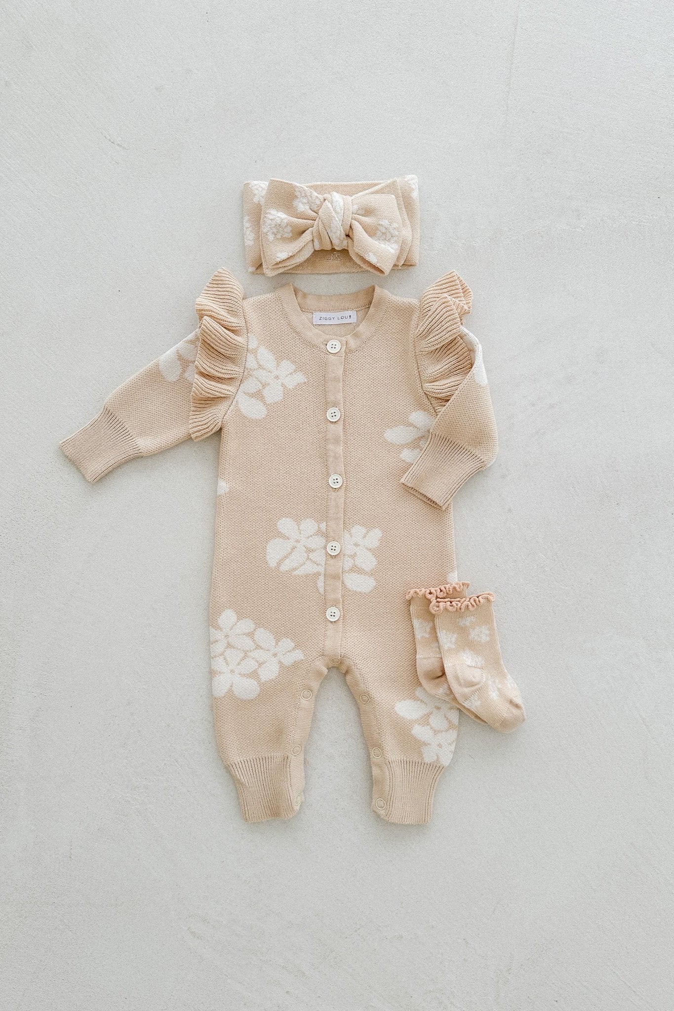 A beige baby onesie with white floral patterns lies on a light background. The ZIGGY LOU outfit features ruffled sleeves and front buttons. Nearby, a matching headband with a bow and a pair of "Socks Banksy," also beige with white floral patterns, are placed, all part of their charming Spring Collections.