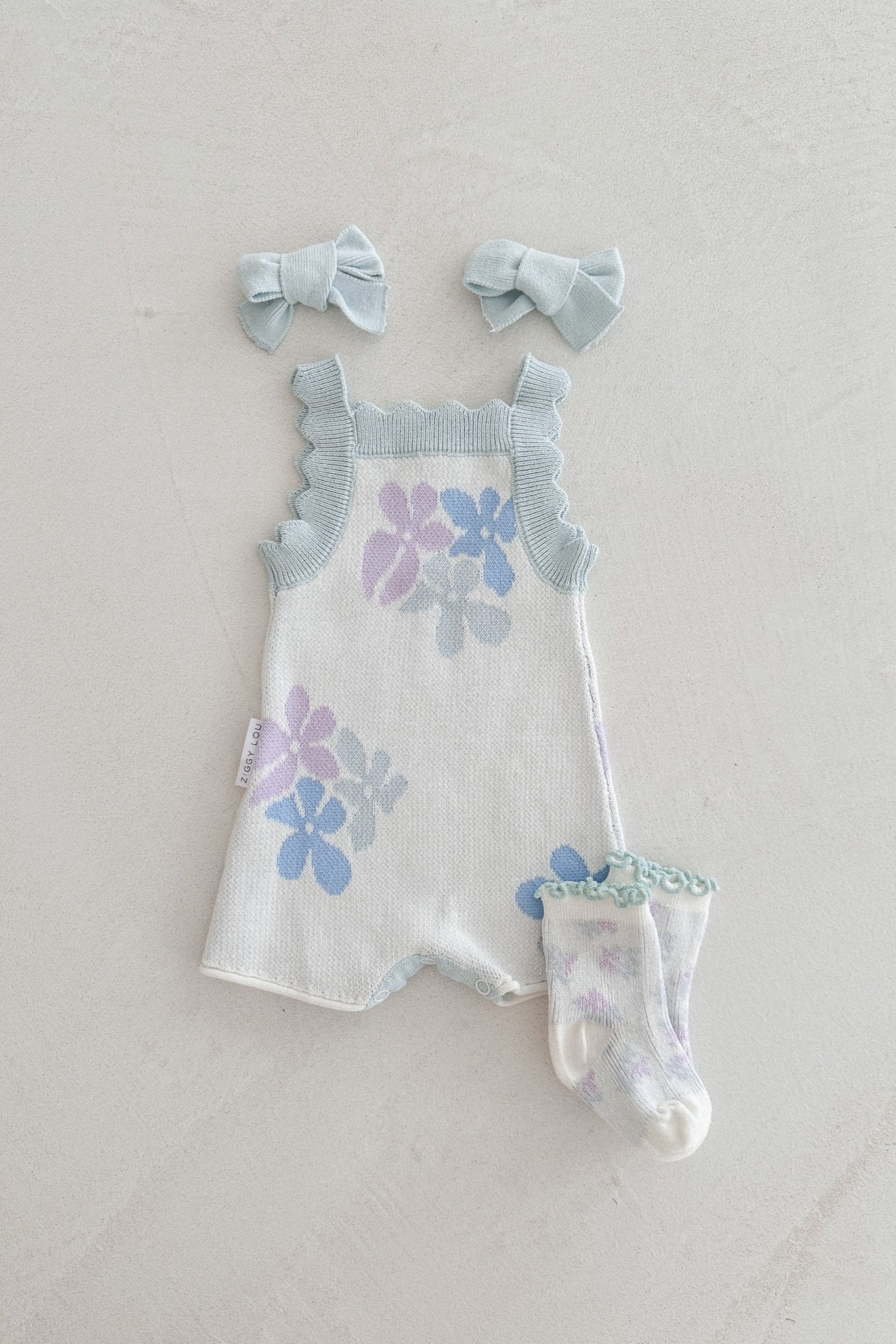 The Playsuit Alaska by ZIGGY LOU, a knitted baby romper adorned with light blue and purple flowers, is laid out on a light background. Matching light blue hair bows and floral socks complete the outfit. This 100% cotton romper features functional press snaps and ruffled straps, adding a cute touch to this adorable set.