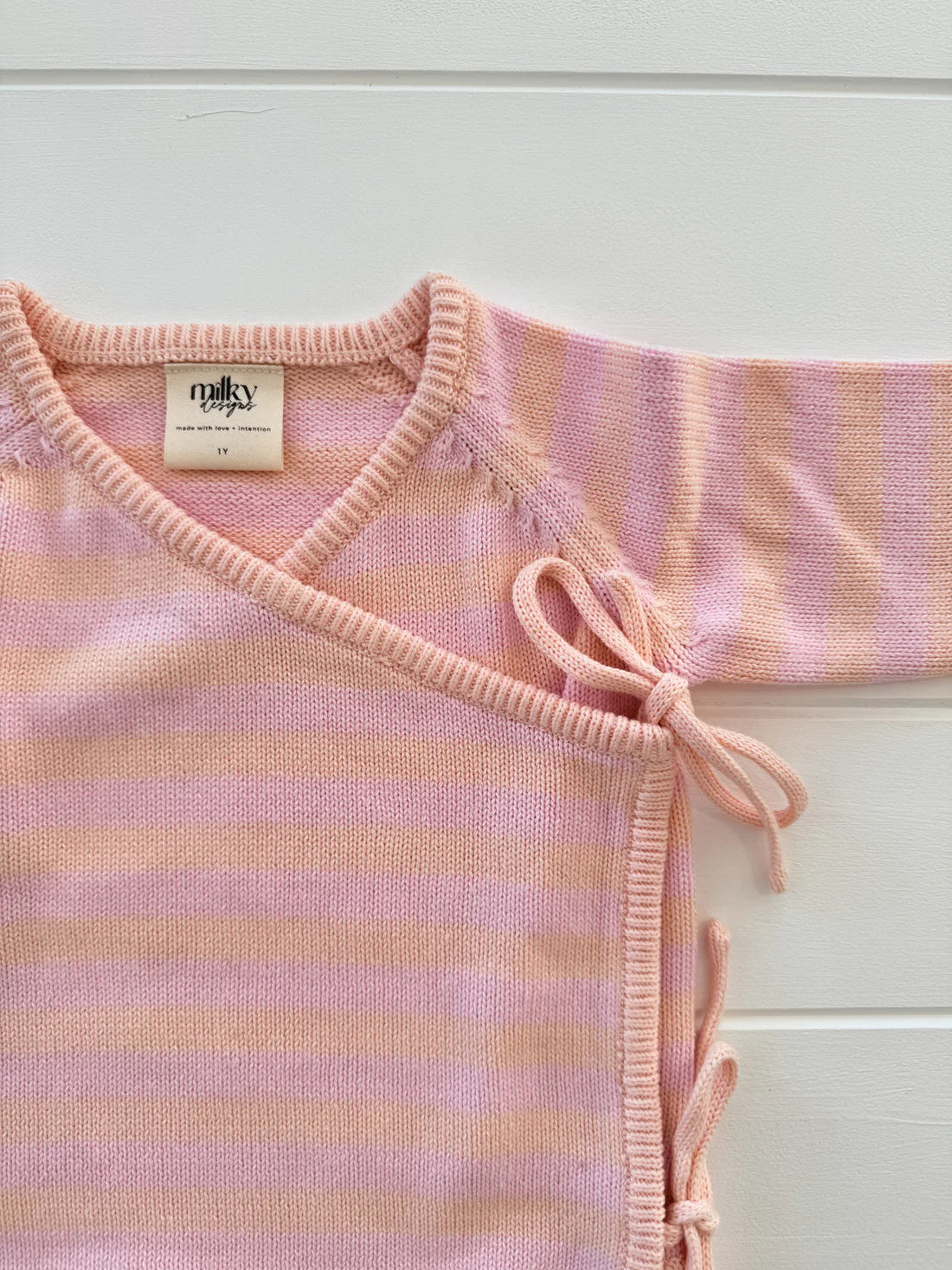 The Essential Wrap Romper Pink/Peach by MILKY DESIGNS is showcased against a white background. This 100% cotton romper boasts a pink and white striped pattern on soft knit fabric. It features an overlapping front secured with small side ties for closure, ensuring maximum comfort for your baby. The label inside reads "malu" and indicates the size "3M.