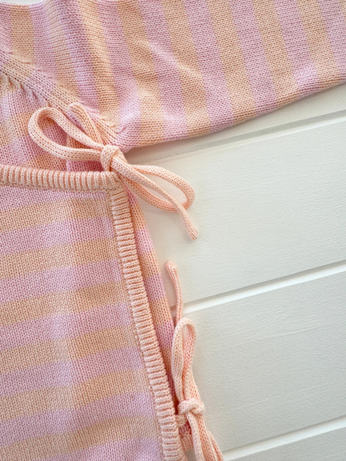 Close-up of the MILKY DESIGNS Essential Wrap Romper in Pink/Peach, showcasing its knitted fabric with pink and beige horizontal stripes. The romper's charm is enhanced by neatly tied bows made from the same 100% cotton material, making it perfect for baby clothing. The background features a white paneled surface, adding to the sense of comfort and coziness.