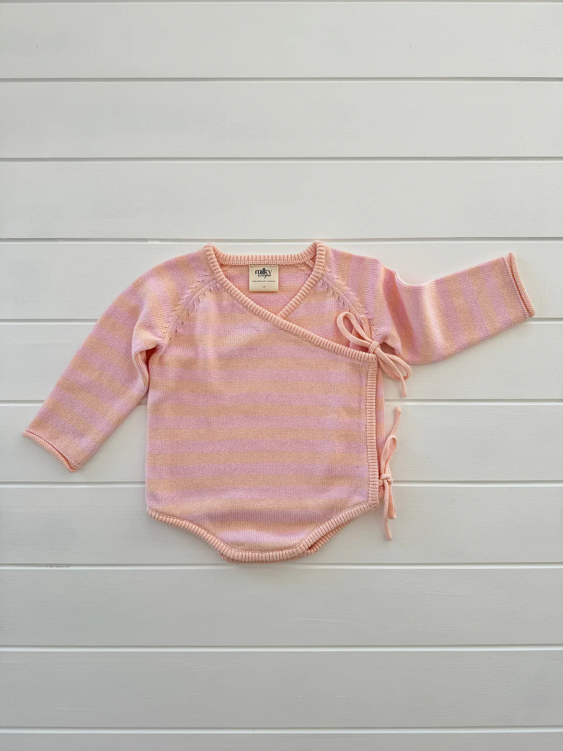 The Essential Wrap Romper Pink/Peach from MILKY DESIGNS, a pink and light purple striped baby kimono sweater made from 100% cotton, is laid flat on a white wooden surface. This comfortable romper features long sleeves, a V-neckline, and side ties for closure.