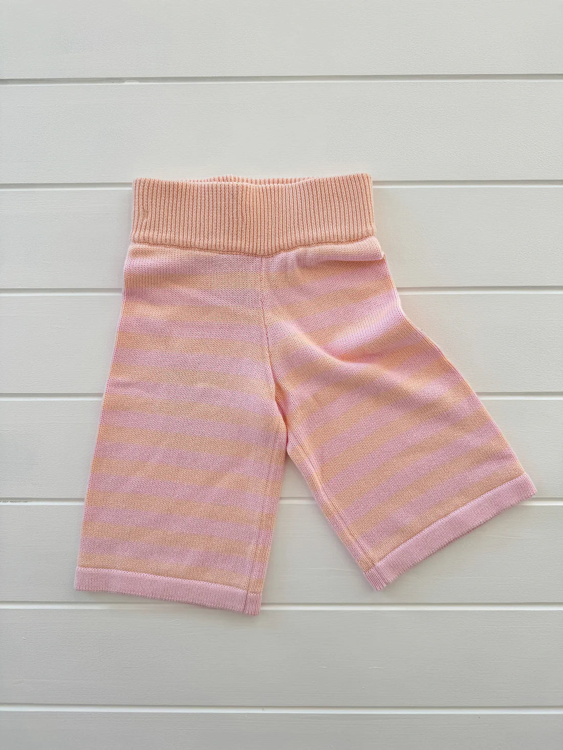 The MILKY DESIGNS Staple Pant Pink/Peach, a pair of children's shorts with pink and orange stripes, is displayed on a white wooden surface. Made from 100% cotton knit, these shorts feature a ribbed waistband and are laid flat to showcase their pattern and design. Available in sizes from 0-4 years, they ensure a perfect fit for your little one.