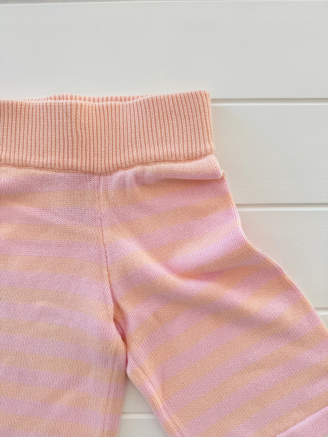 Close-up of the waistband and part of one leg of the MILKY DESIGNS Staple Pant Pink/Peach. These 100% cotton knit pants feature alternating horizontal stripes in light pink and peach colors and have a ribbed elastic waistband. Available in sizes ranging from 0-4 years, they are displayed against a white background with horizontal paneling.