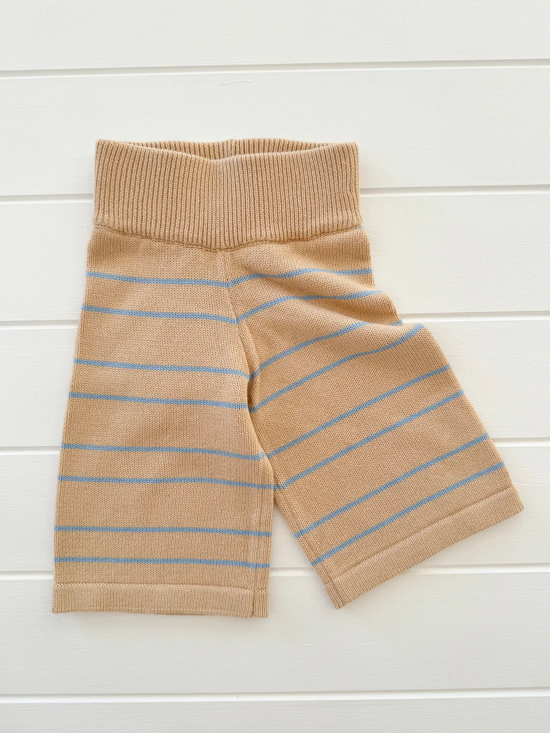 The Staple Pant Blue Stripe by MILKY DESIGNS is displayed flat against a white wooden background. These beige knitted shorts with light blue horizontal stripes are crafted from 100% cotton knit, featuring a wide ribbed elastic waistband and providing a comfortable 3/4 fit.