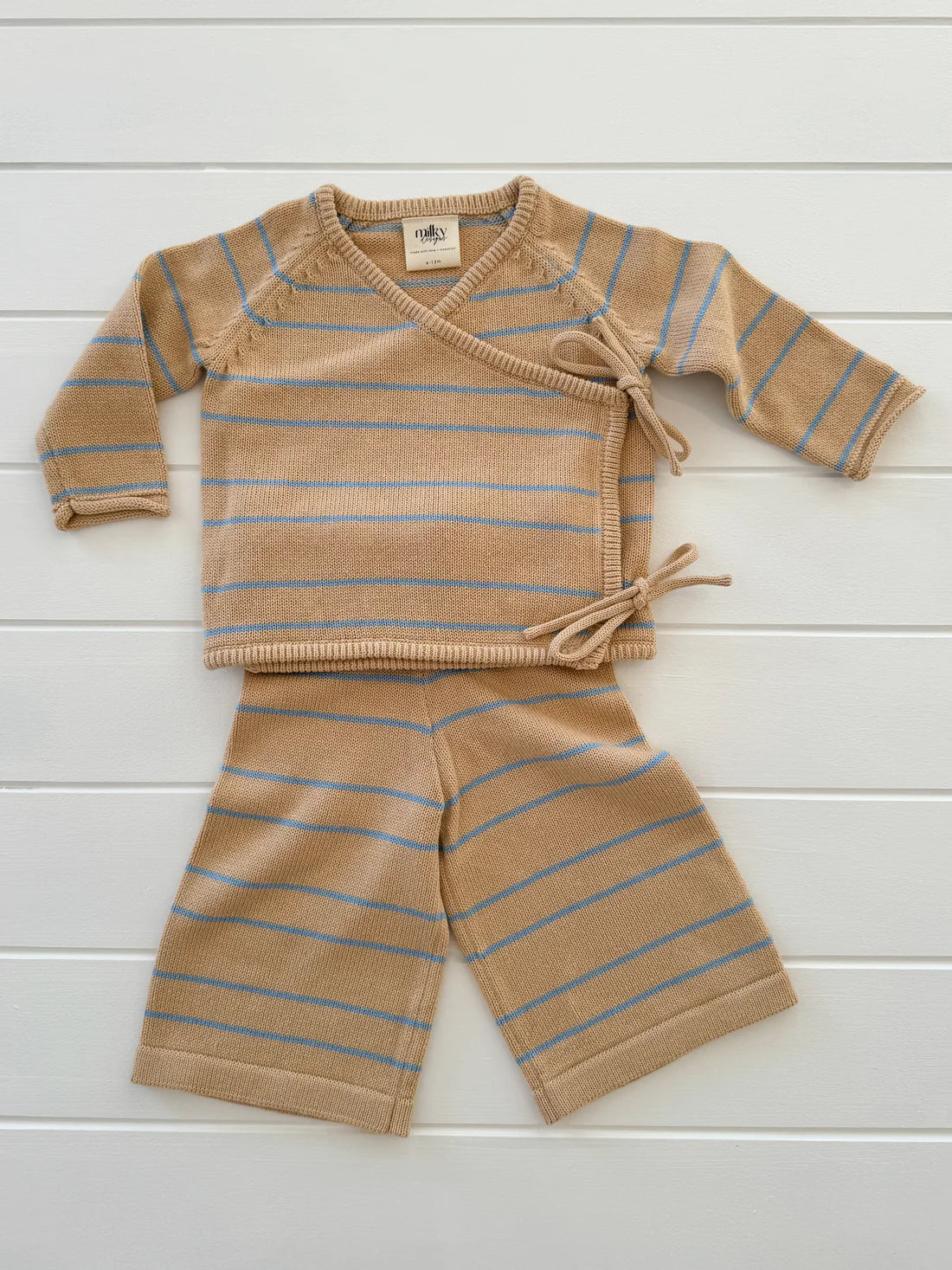 A tan-colored baby outfit called the Staple Pant Blue Stripe by MILKY DESIGNS, featuring a long-sleeve wrap sweater with side ties and matching wide-legged pants with an elastic waistband, laid out on a white wooden surface.