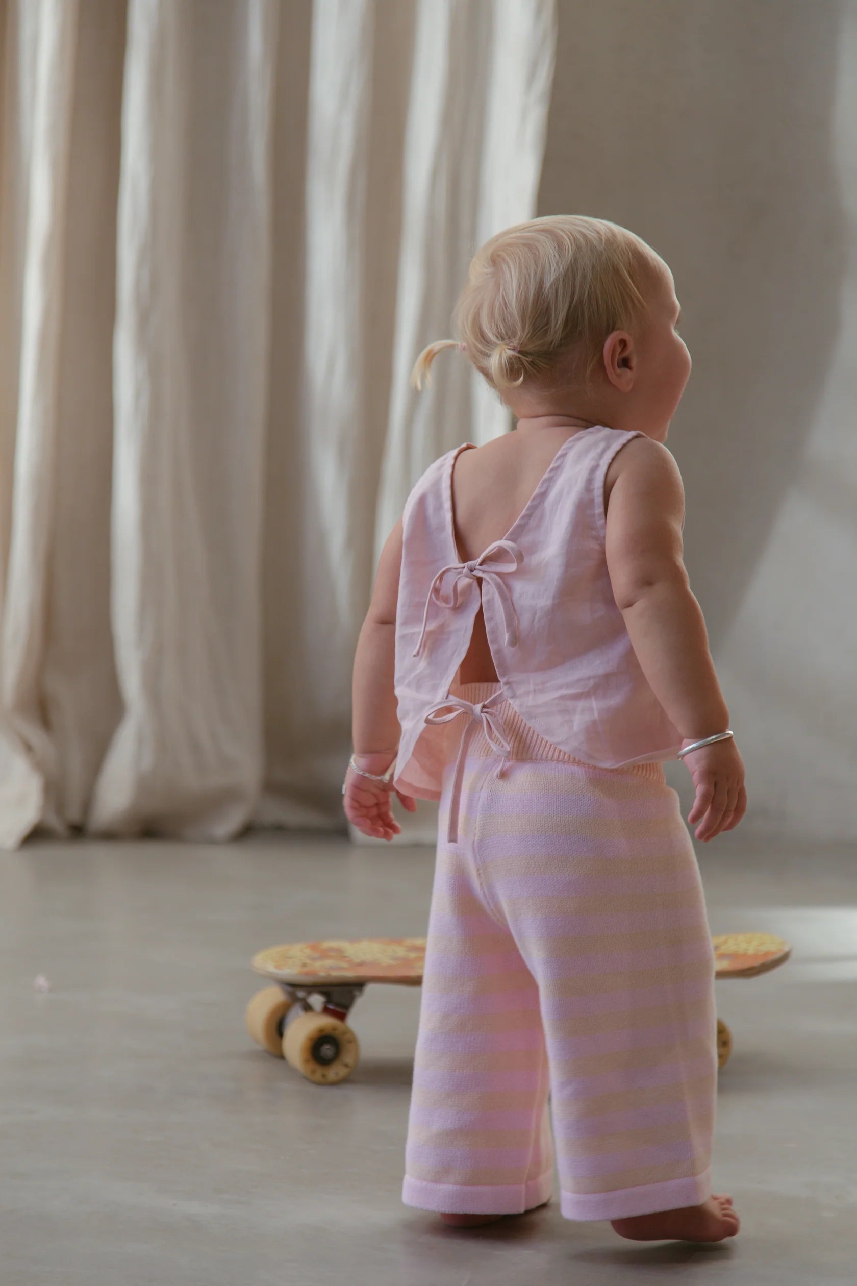 A toddler with blond hair, dressed in the Staple Pant Pink/Peach from MILKY DESIGNS, stands indoors on a smooth floor. A skateboard is visible on the floor behind the child. Light cream-colored curtains hang in the background.