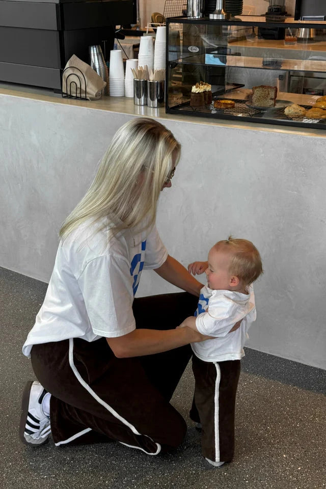 A woman with long blonde hair and wearing glasses kneels down while holding her young child. They are both dressed in matching white T-shirts and Piper Racer Pants in Dark Chocolate, featuring a relaxed fit with an elasticated waistband from the brand TINY TROVE. They are in a modern café with a counter displaying pastries in the background.