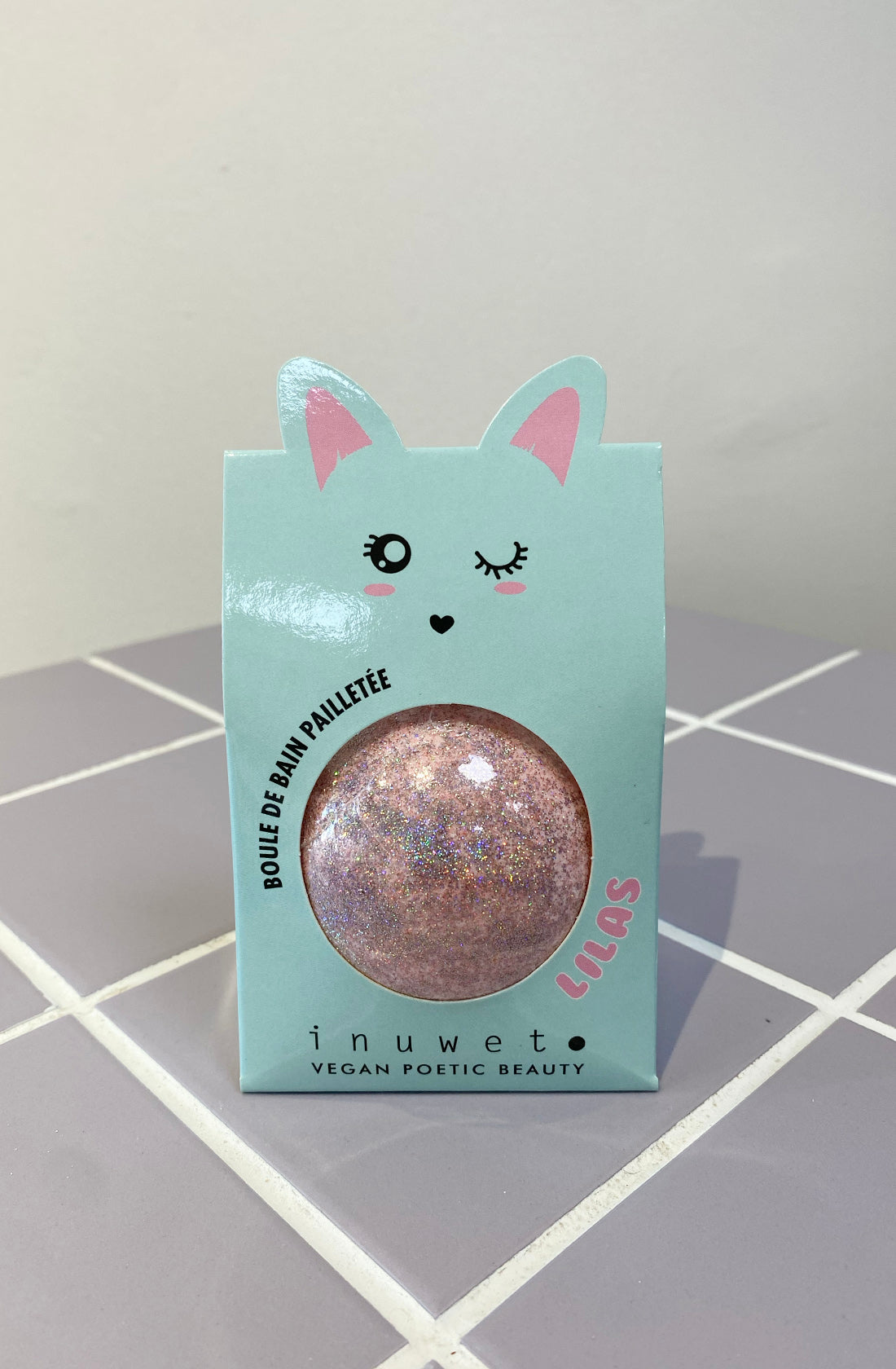 A pastel blue package adorned with cat ears and a winking face holds the Glitter Fizzer Bath Bomb in lilac/pink. The text on the package reads "Boule de Bain Pailletée," "INUWET," and "VEGAN POETIC BEAUTY." With its lilac scent and vegan formula, it sits beautifully against white and light gray tiles.