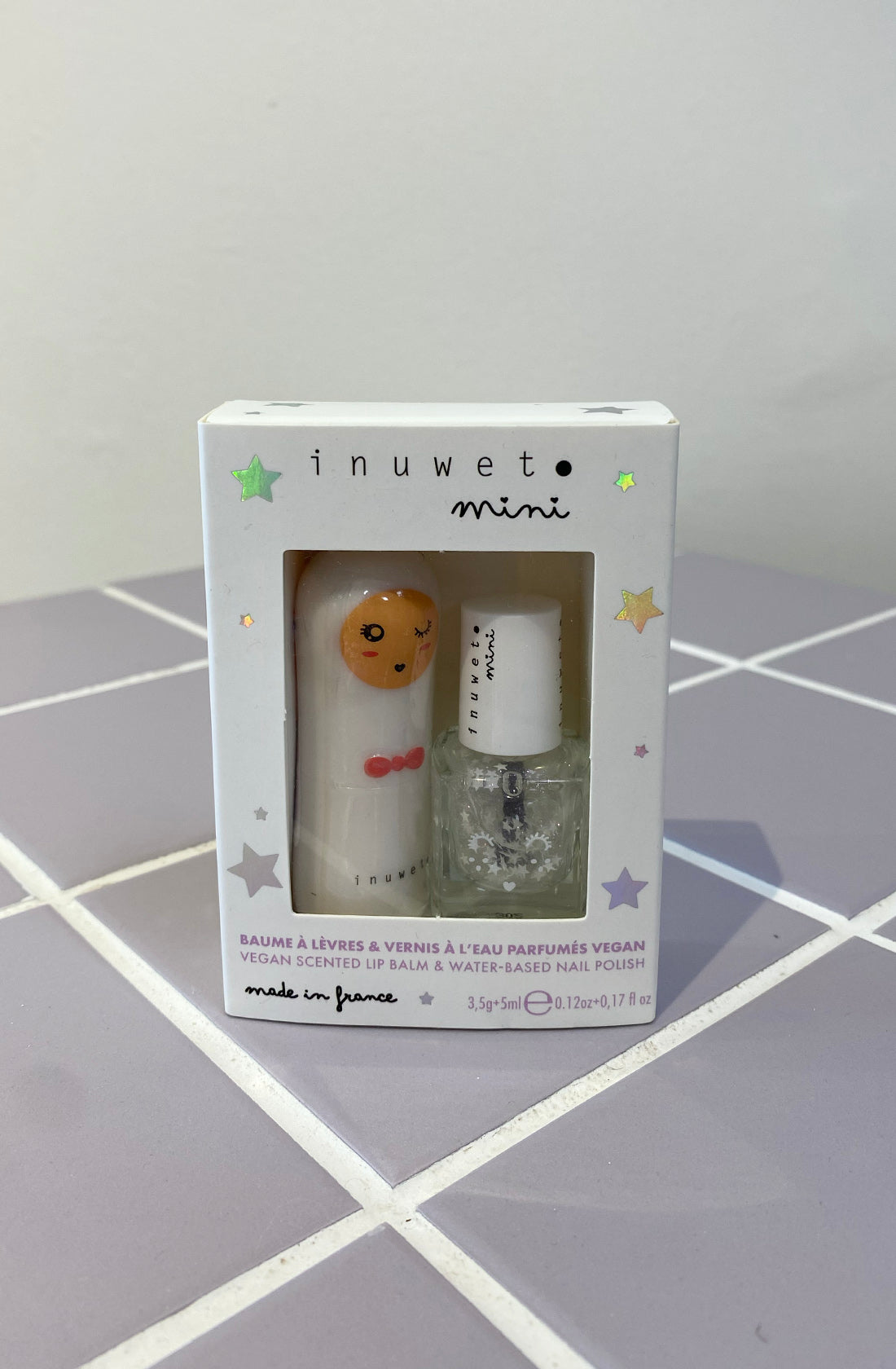 A boxed set of INUWET's Lip Balm & Nail Polish Duo White featuring a vegan cotton candy-scented lip balm and a water-based kids nail polish on a light purple tile surface. The white box has a cutout displaying the products, decorated with stars and text indicating it is made in France.