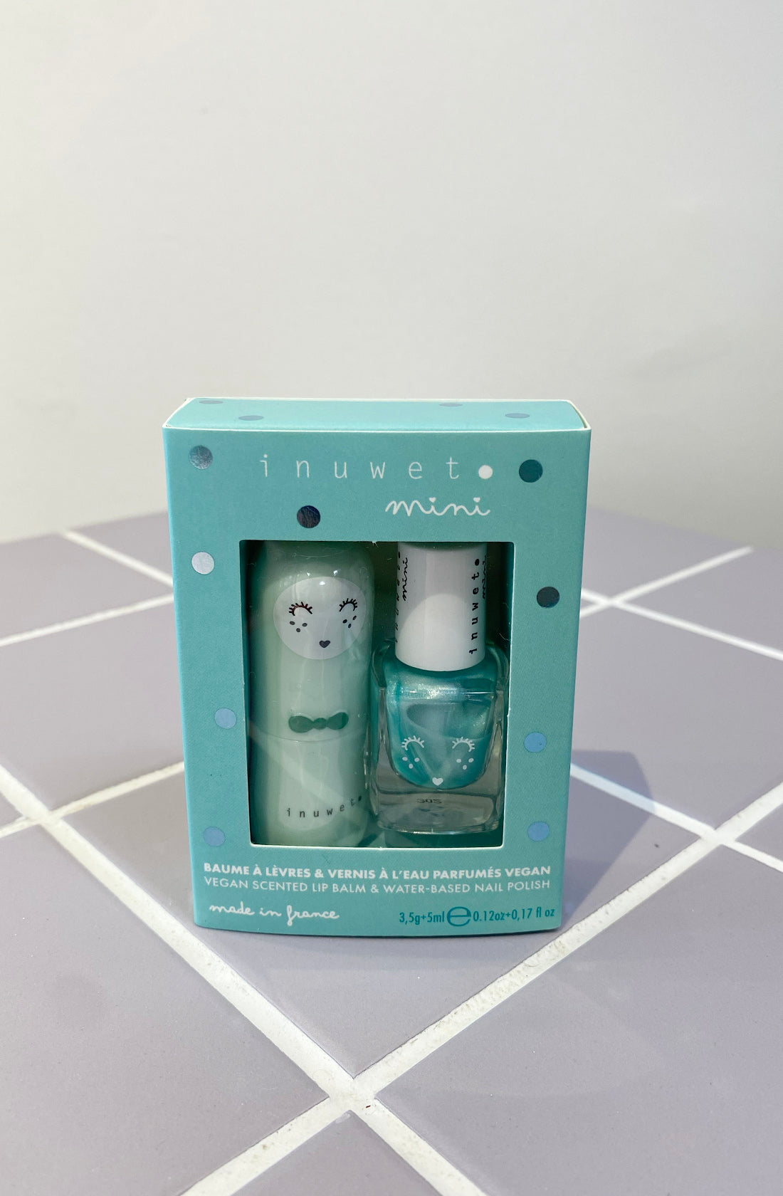 On a tiled surface lies the Lip Balm & Nail Polish Duo Aqua from INUWET, featuring a vegan scented lip balm and kids nail polish made from natural ingredients. The turquoise box with white polka dots showcases charming illustrations, including a smiling face on the lip balm tube and a bunny on the nail polish bottle. The brand name "INUWET mini" is prominently displayed at the top.