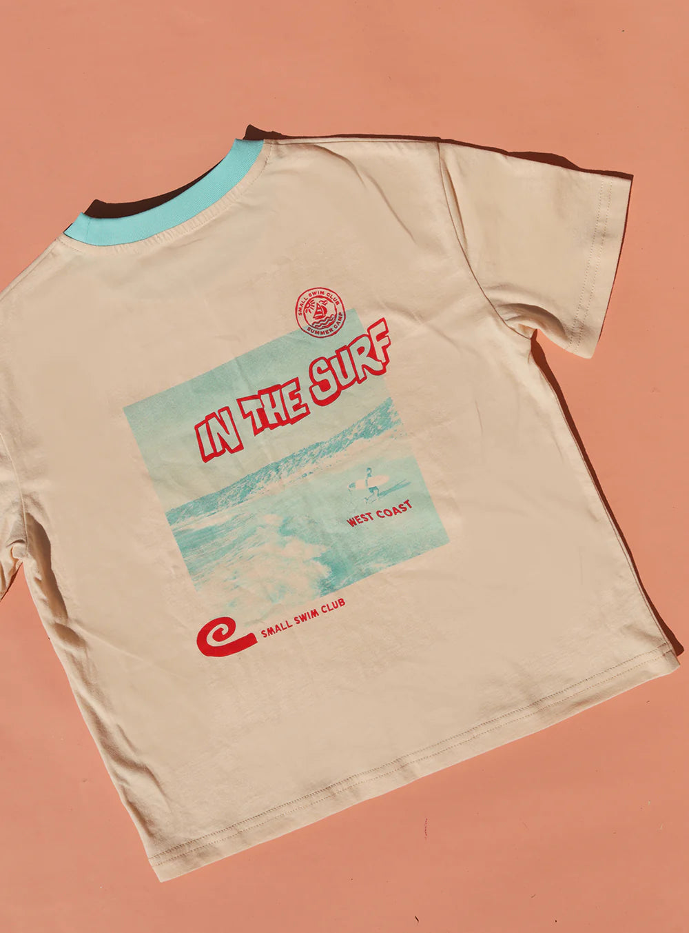 A cream cotton tee from Small Swim Club features a turquoise collar, vintage ocean wave graphics, and the text "In the Surf" and "West Coast." Laid against a warm peach background, it boasts coastal charm.