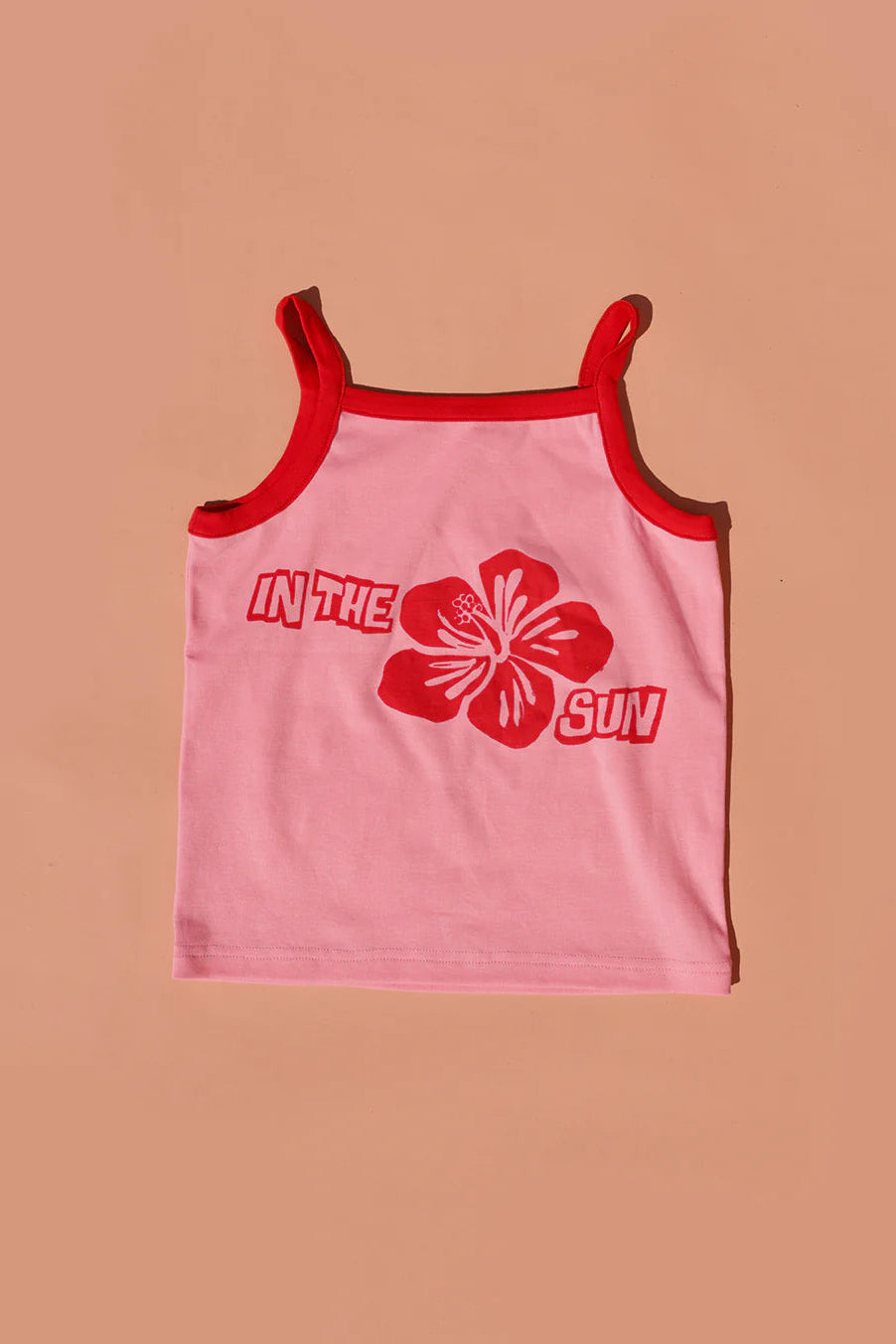 The SMALL SWIM CLUB Hibiscus Singlet Pink/Red showcases a vibrant hibiscus graphic. With red straps and bold "IN THE SUN" text, it beautifully complements the red floral design on a brown background.