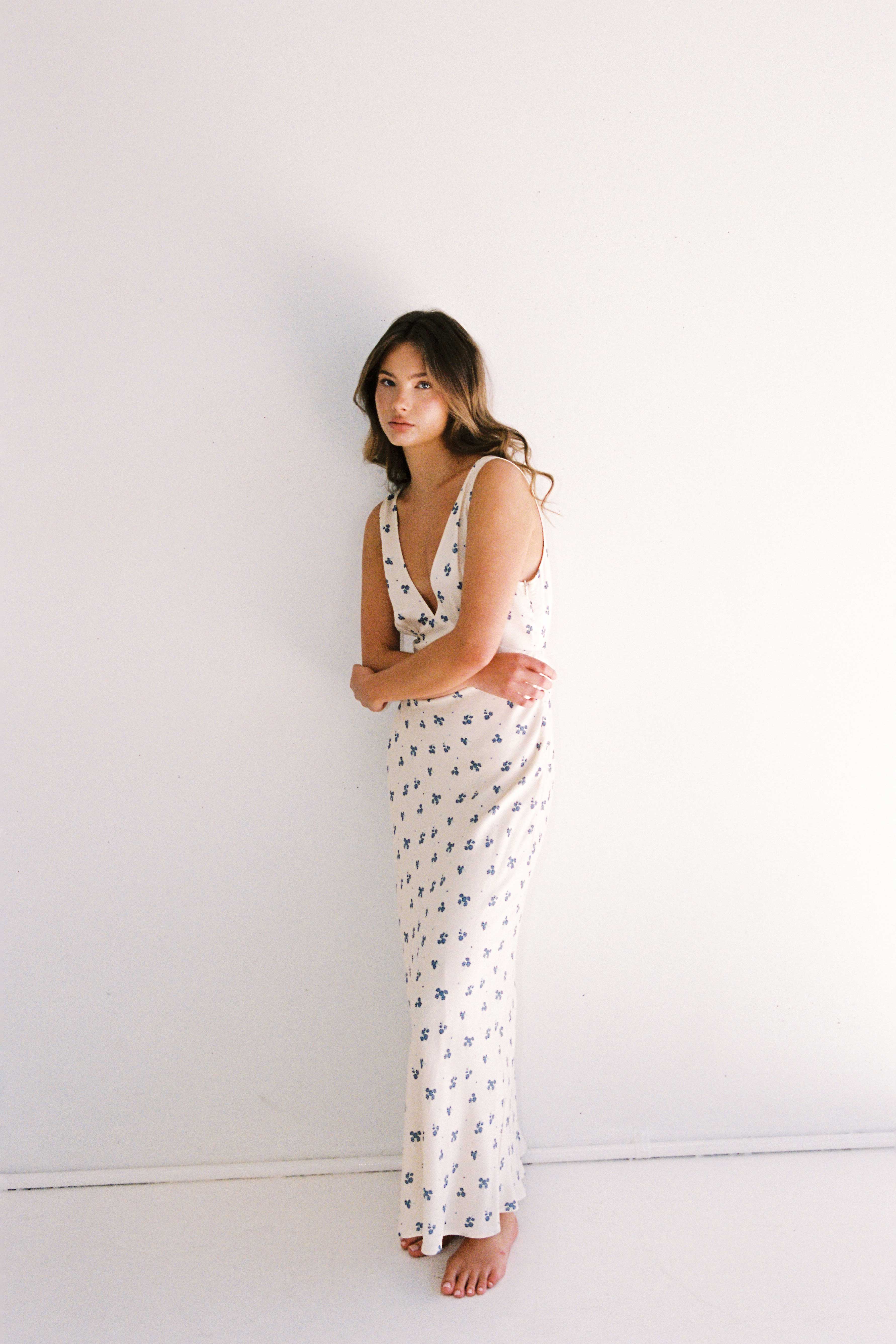 A woman with long hair stands barefoot against a white wall. She wears the "Isabella Midi Dress Ditsy Floral" by JUNI, a sleeveless, floor-length dress adorned with small, dark floral patterns. The ethically made dress features a deep V-neck as she poses with her hands together, looking towards the camera.