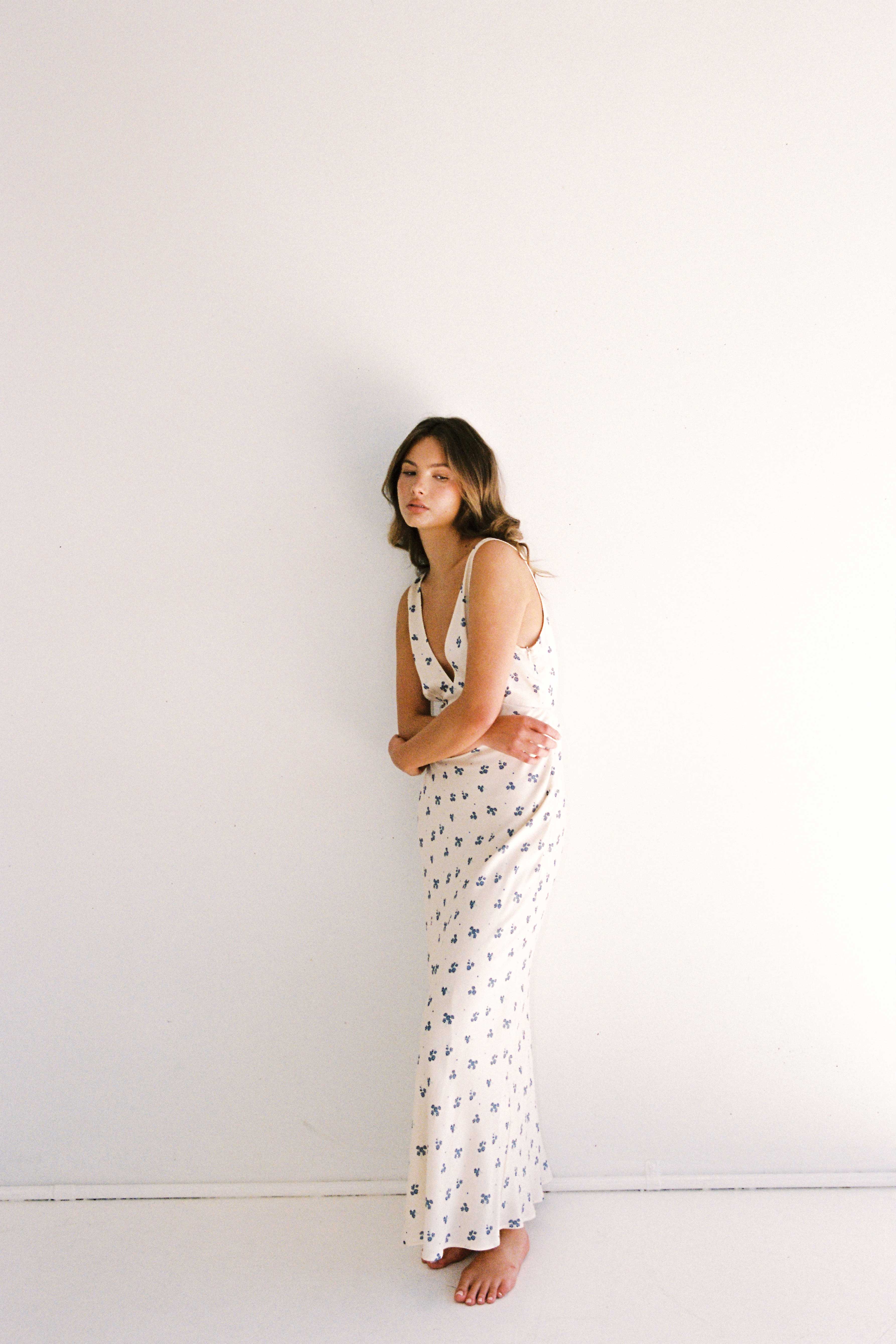 A woman with long brown hair is standing barefoot against a plain white wall. She is wearing the Isabella Midi Dress Ditsy Floral by JUNI, a white sleeveless dress with a v-neckline adorned with small blue floral patterns. She has her arms gently crossed and looks calm and composed.