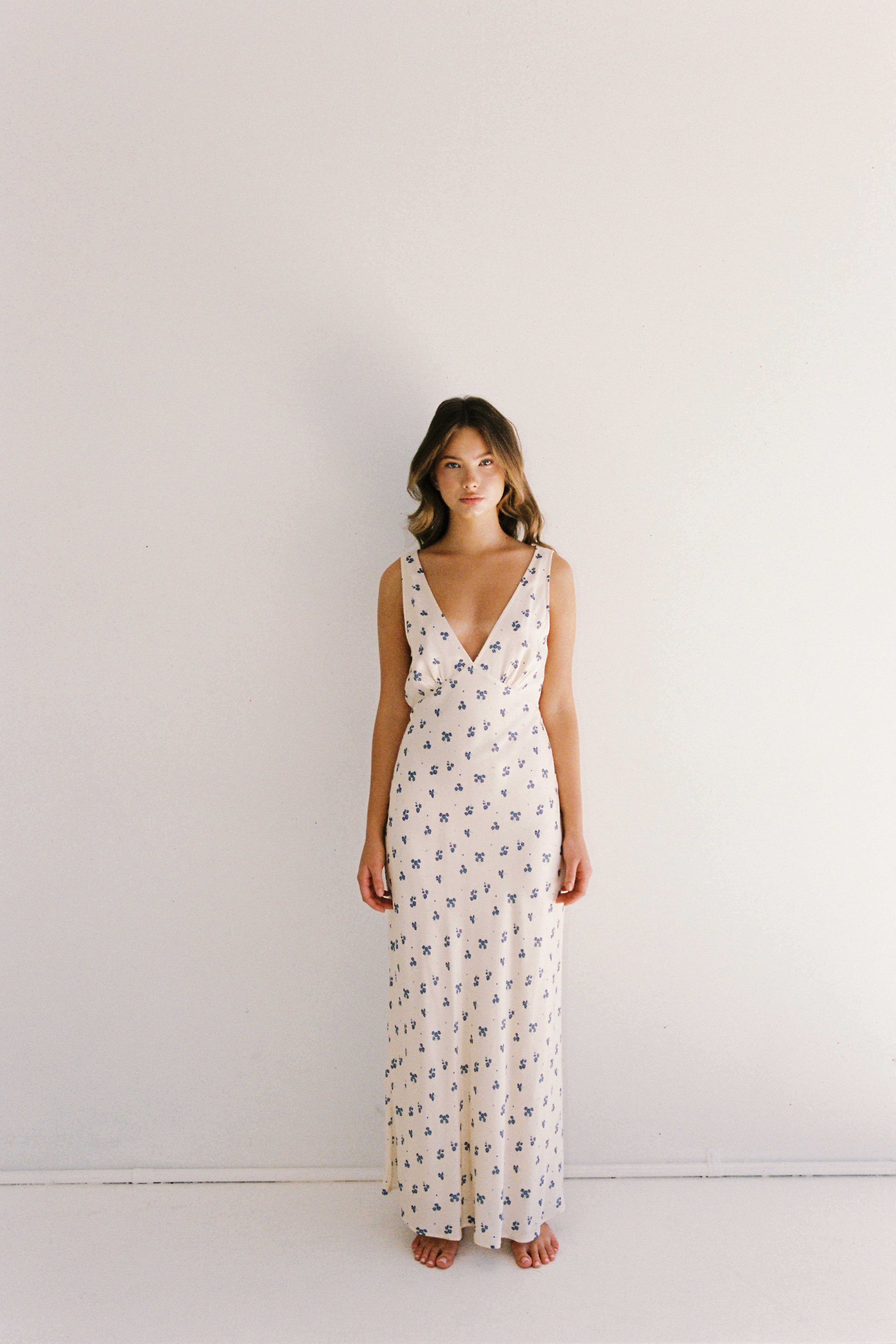 A woman with long, wavy hair stands barefoot against a plain white wall, wearing the ethically made Isabella Midi Dress Ditsy Floral from JUNI. The sleeveless dress features a deep V-neckline and is adorned with a delicate blue floral print. She gazes directly at the camera with a relaxed posture.