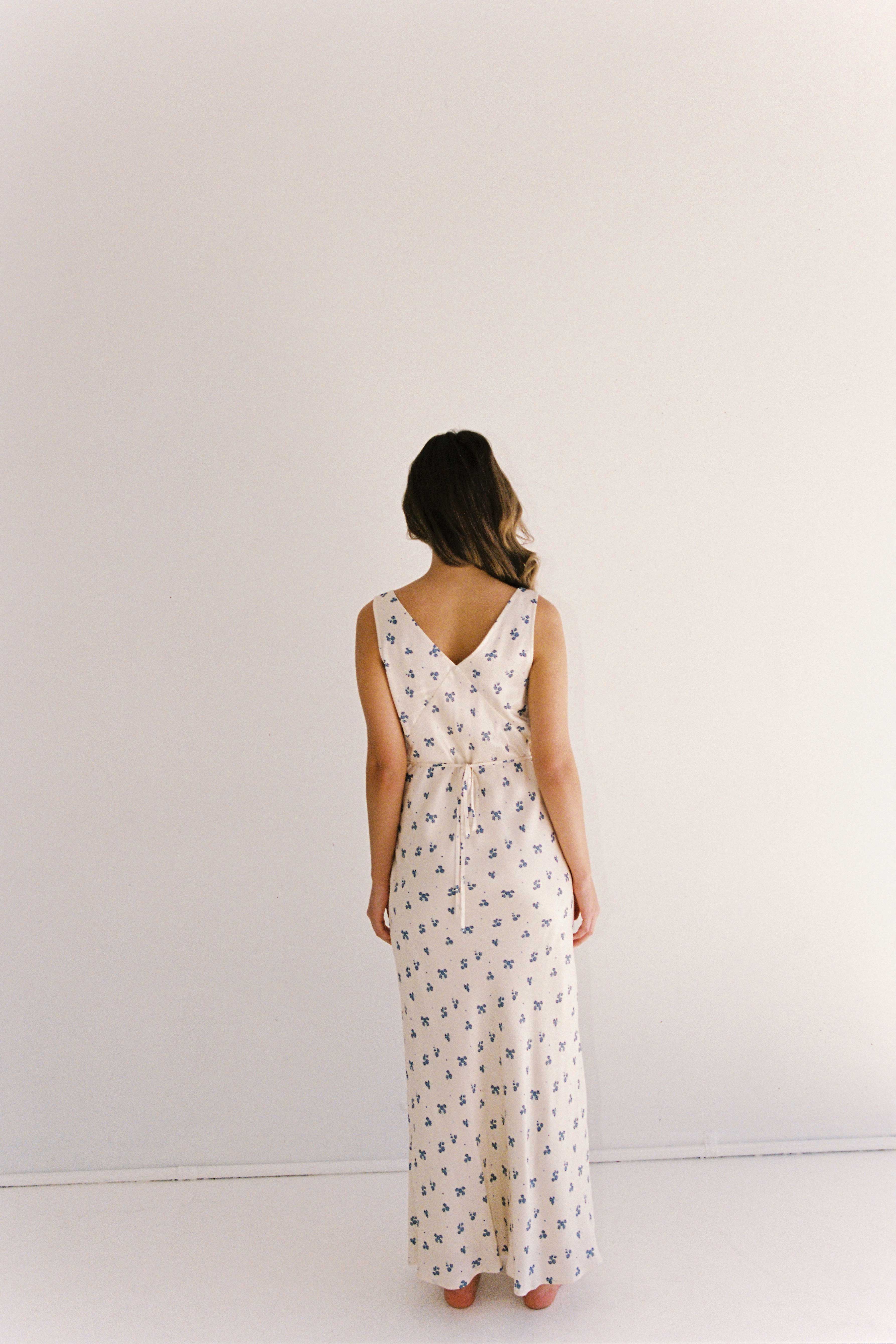 A woman with wavy, medium-length hair stands facing away, barefoot against a plain, light-colored wall. She is wearing the ethically made JUNI Isabella Midi Dress in Ditsy Floral, a sleeveless, ankle-length dress with a small floral pattern tied at the waist with a fabric belt.