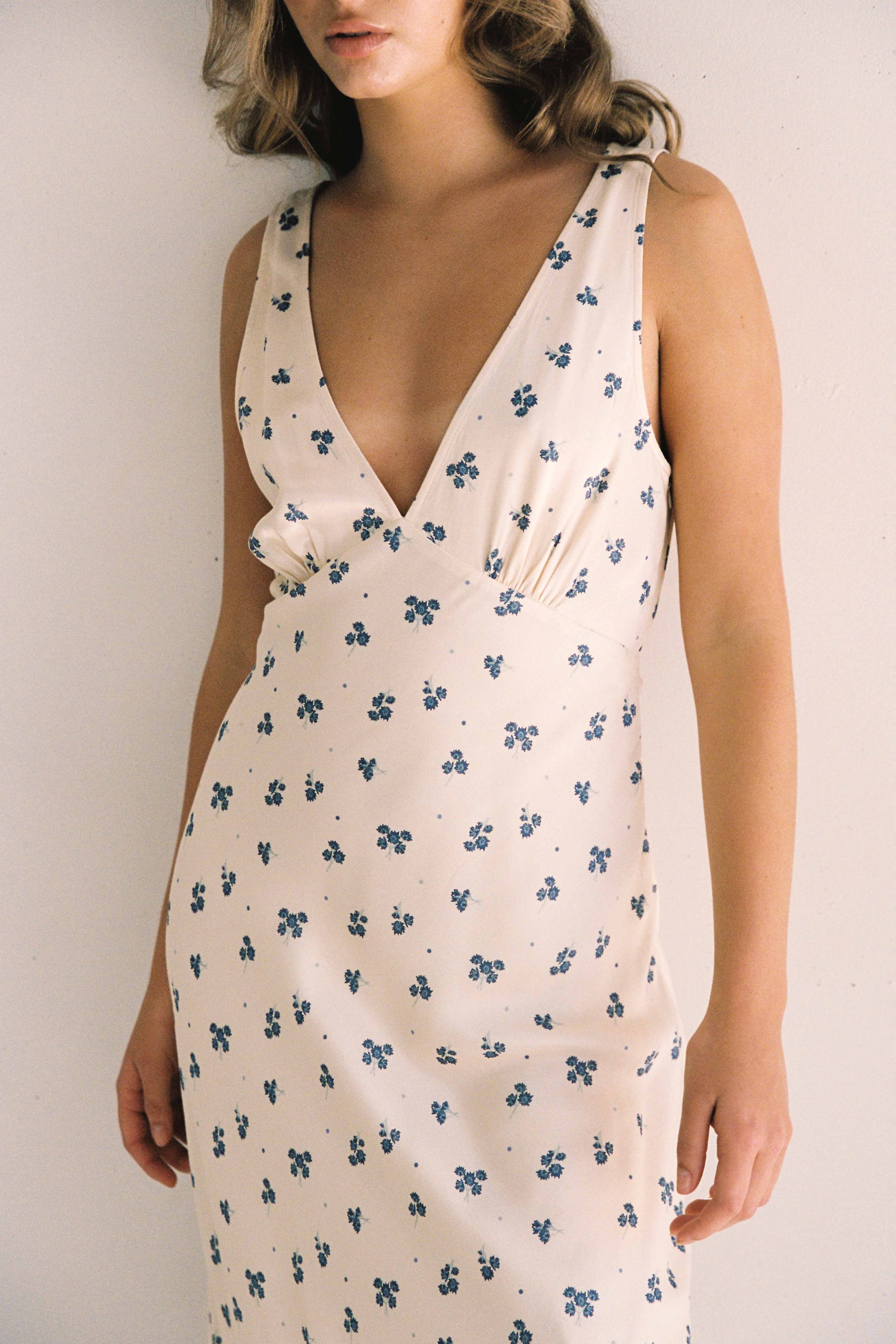 A person wearing the Isabella Midi Dress Ditsy Floral from JUNI. This cream-colored, ethically made dress features a bias cut and a deep V-neckline, adorned with small blue floral designs. The person's head is partially out of frame as they stand against a plain, light-colored background.