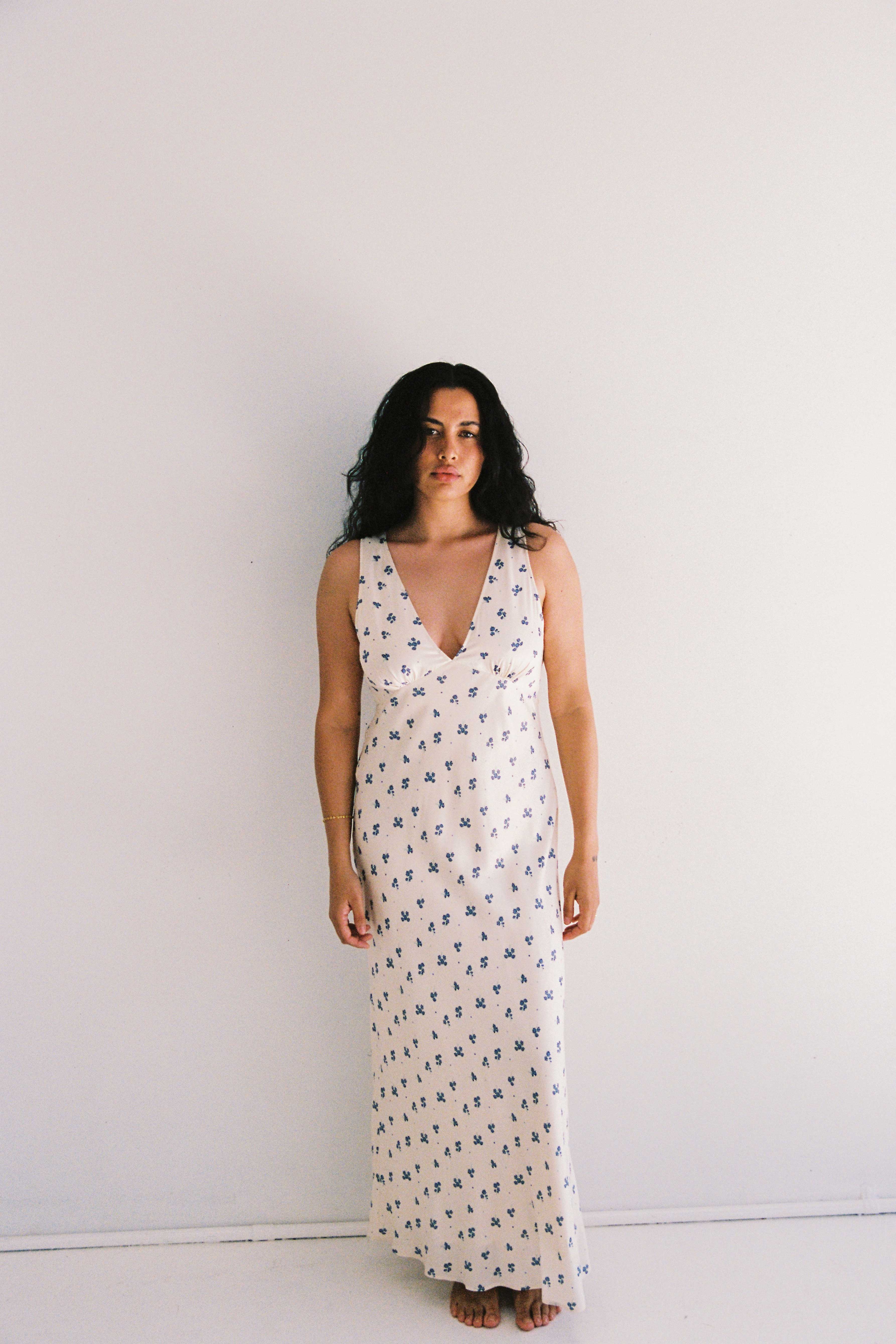 A woman with long, dark, wavy hair stands barefoot against a plain white wall. She is wearing the Isabella Midi Dress Ditsy Floral by JUNI, which is an ethically made, sleeveless dress with a plunging v neckline and blue floral pattern.