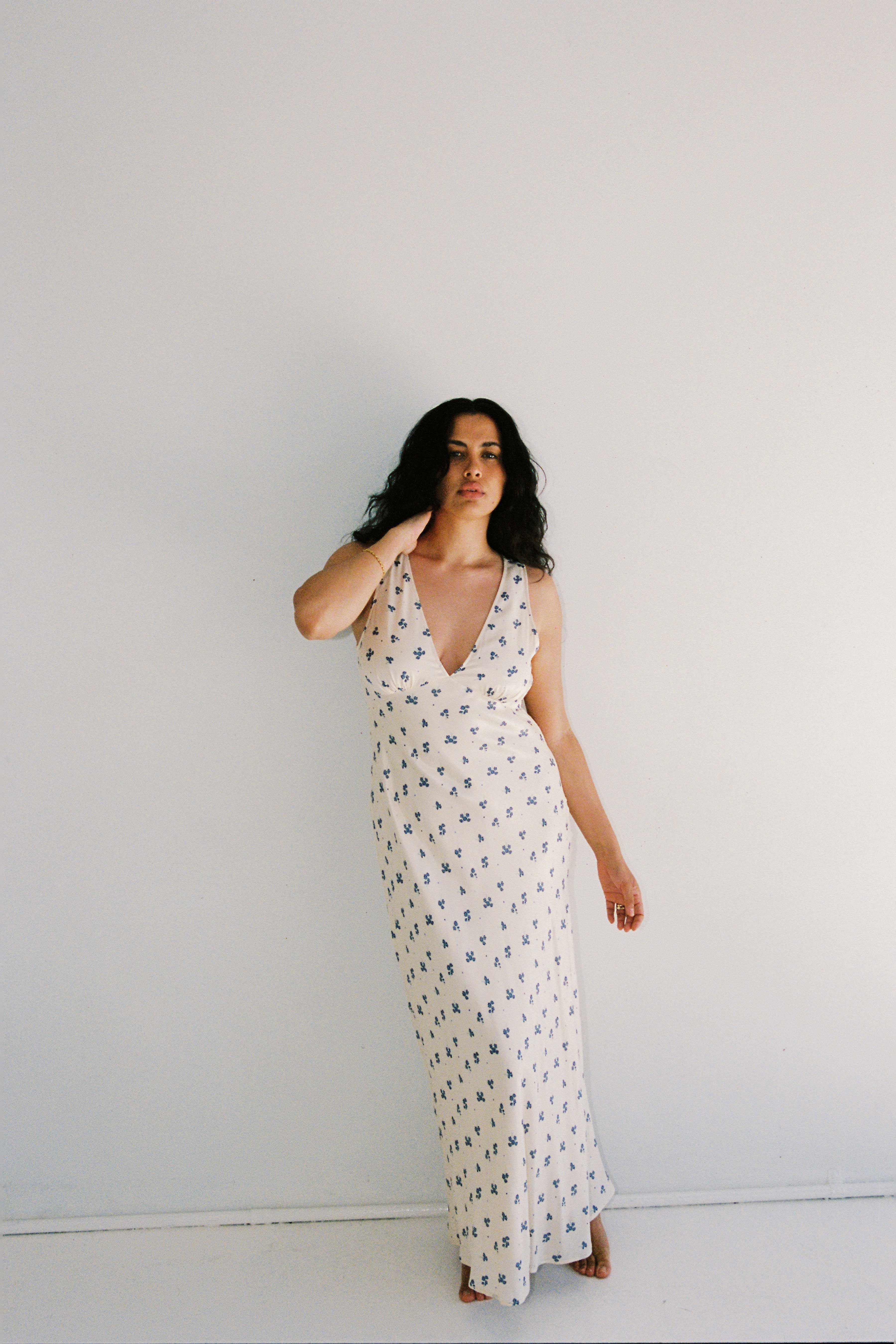 A woman with long, dark hair stands against a plain white background. She wears the "Isabella Midi Dress Ditsy Floral" by JUNI, an ethically made dress adorned with a small black floral pattern. The sleeveless dress features a deep V-neckline and a relaxed fit. With one hand gently touching her hair, she exudes effortless elegance.