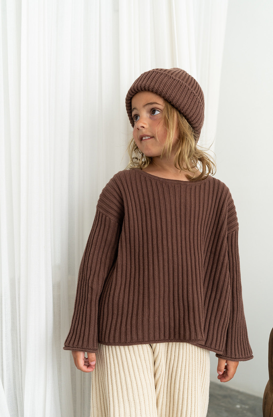 A young kid wearing the essential knit jumper in cocoa with matching beanie.