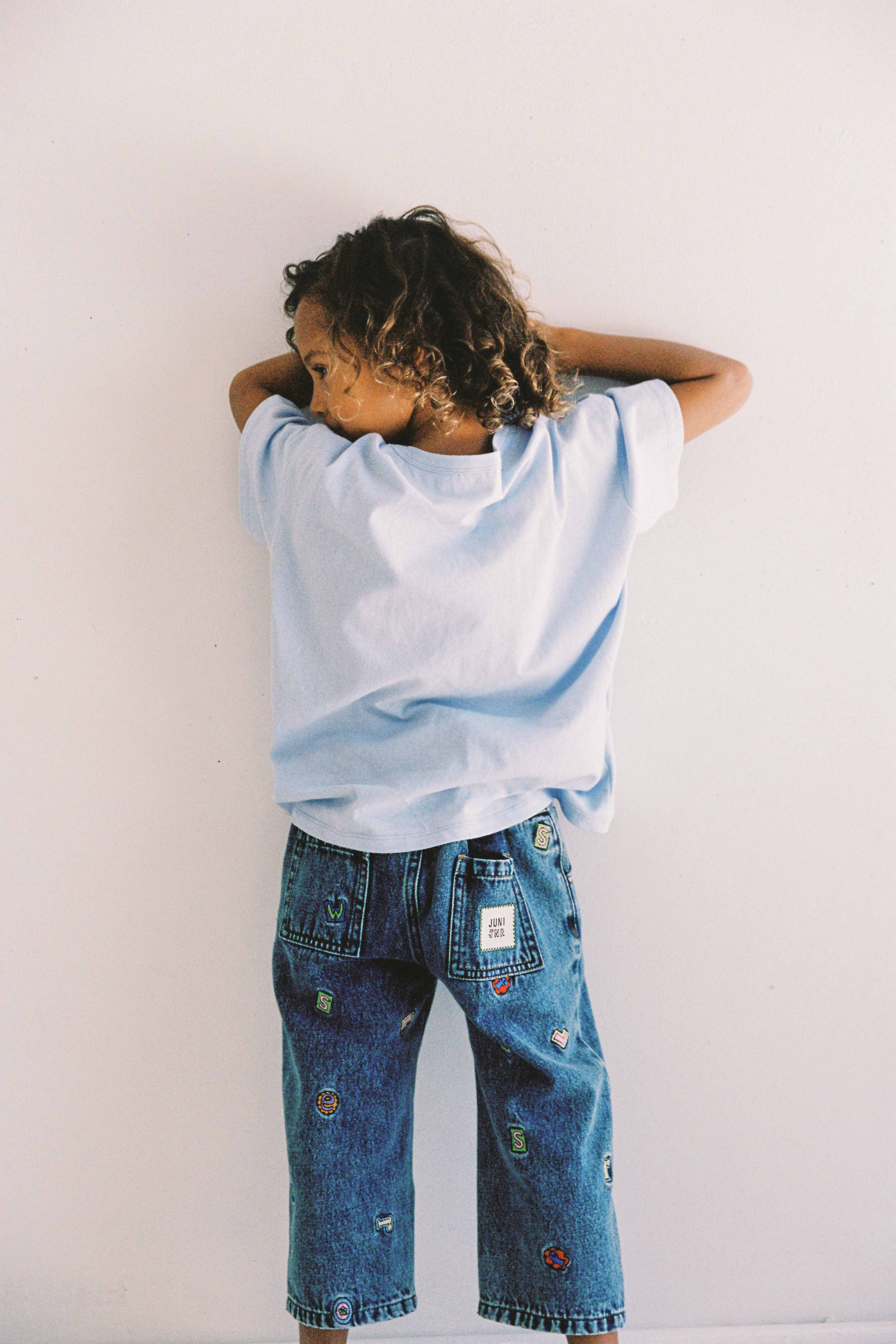 A young child with curly hair stands against a white wall, facing away. They are wearing an oversized light blue t-shirt and JUNI JUNIOR's Alphabet Denim Jean, which features an elastic waistband adorned with colorful patches and embroidery designs. The child has their arms raised and resting on the wall.