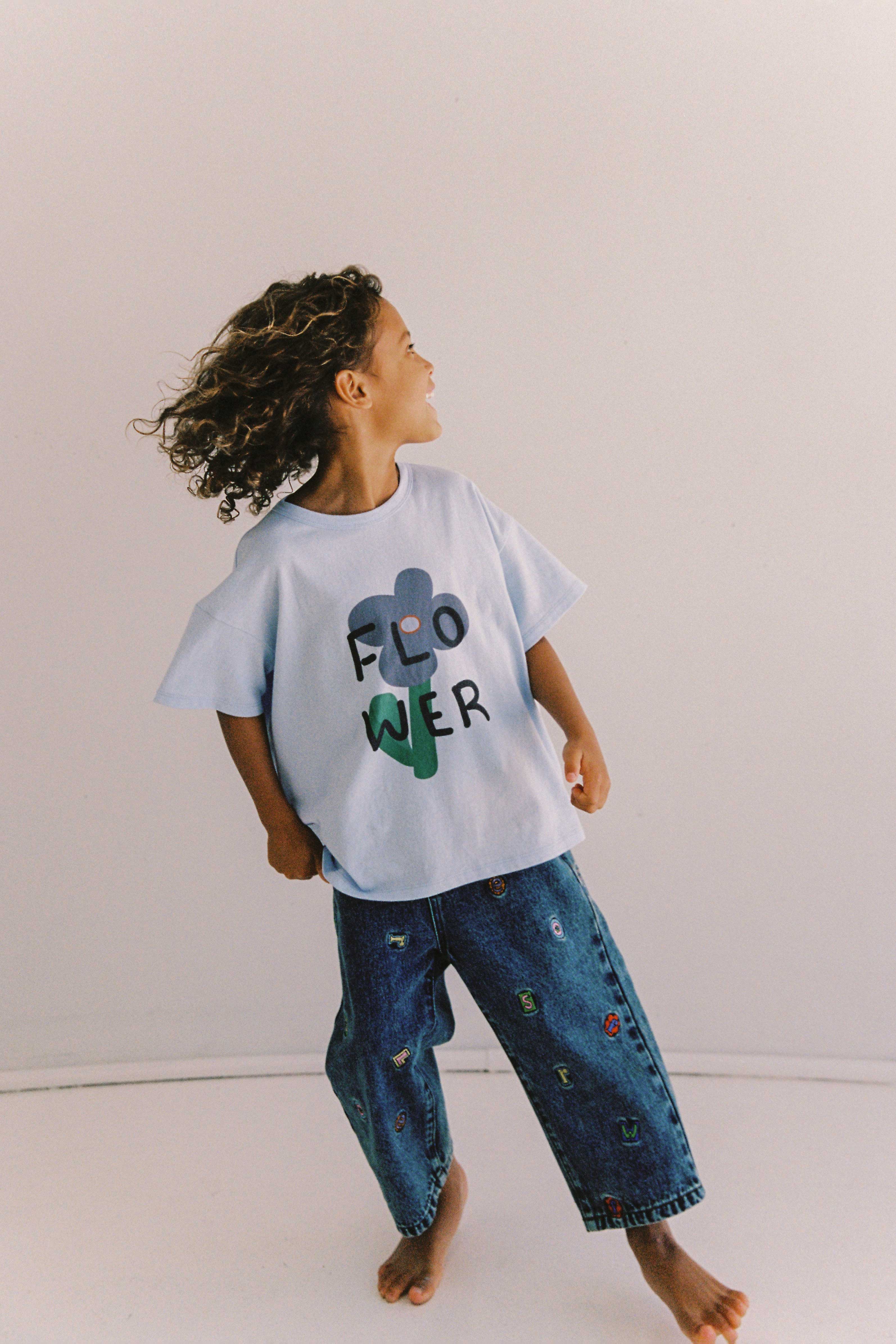 A child with curly hair looks to the side while wearing a light blue t-shirt with the word "FLOWER" printed on it in colorful letters. The child also wears JUNI JUNIOR's Alphabet Denim Jean, which are high-waisted, wide-legged blue jeans featuring a small embroidered pattern and stands barefoot against a white background.