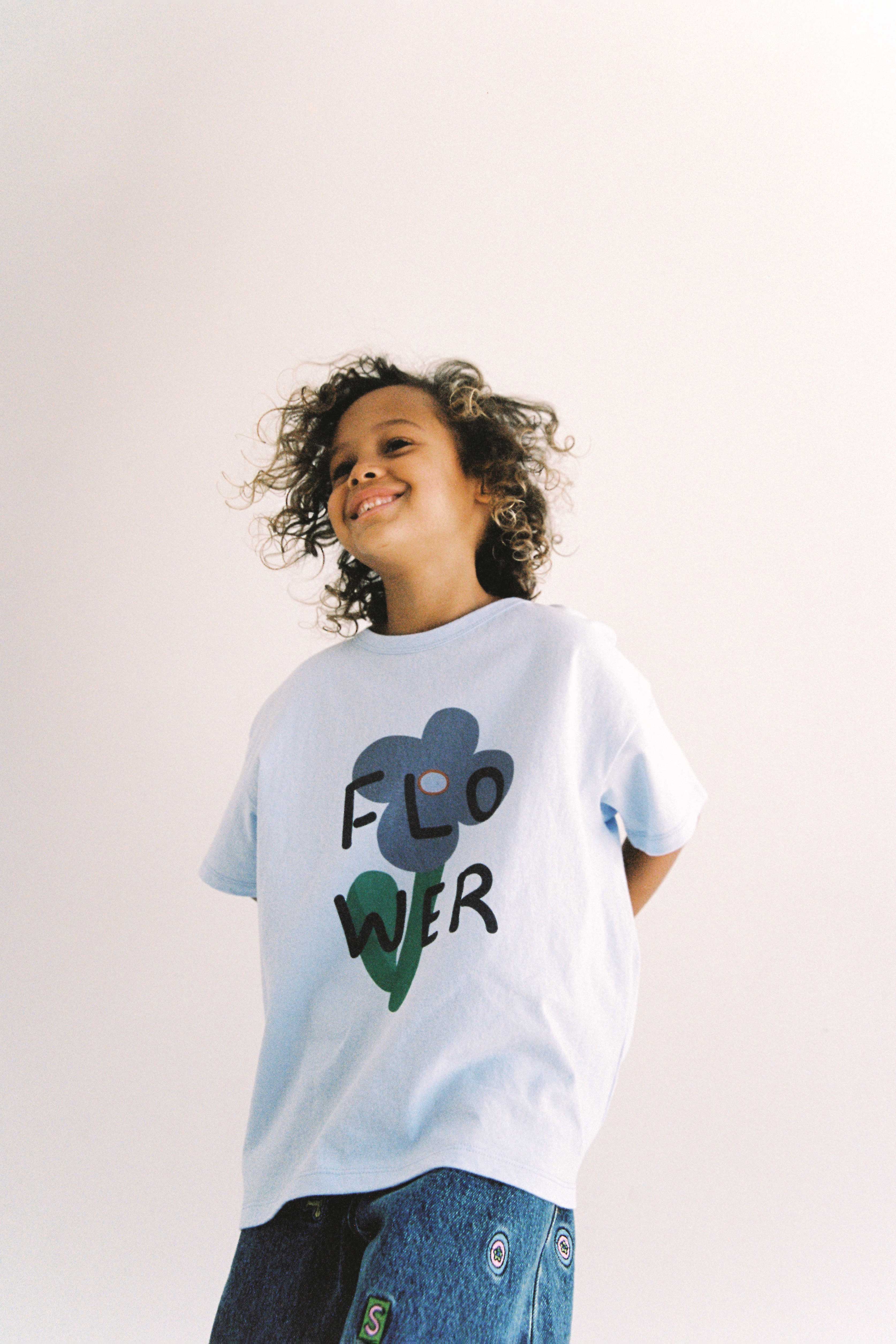 A child with curly hair smiles while wearing the JUNI JUNIOR Junior Bloom Tee Light Blue, made from GOTS certified organic cotton. The shirt features the word "FLOWER" and a graphic of a flower. The child also wears blue jeans with patches, set against a plain white background.