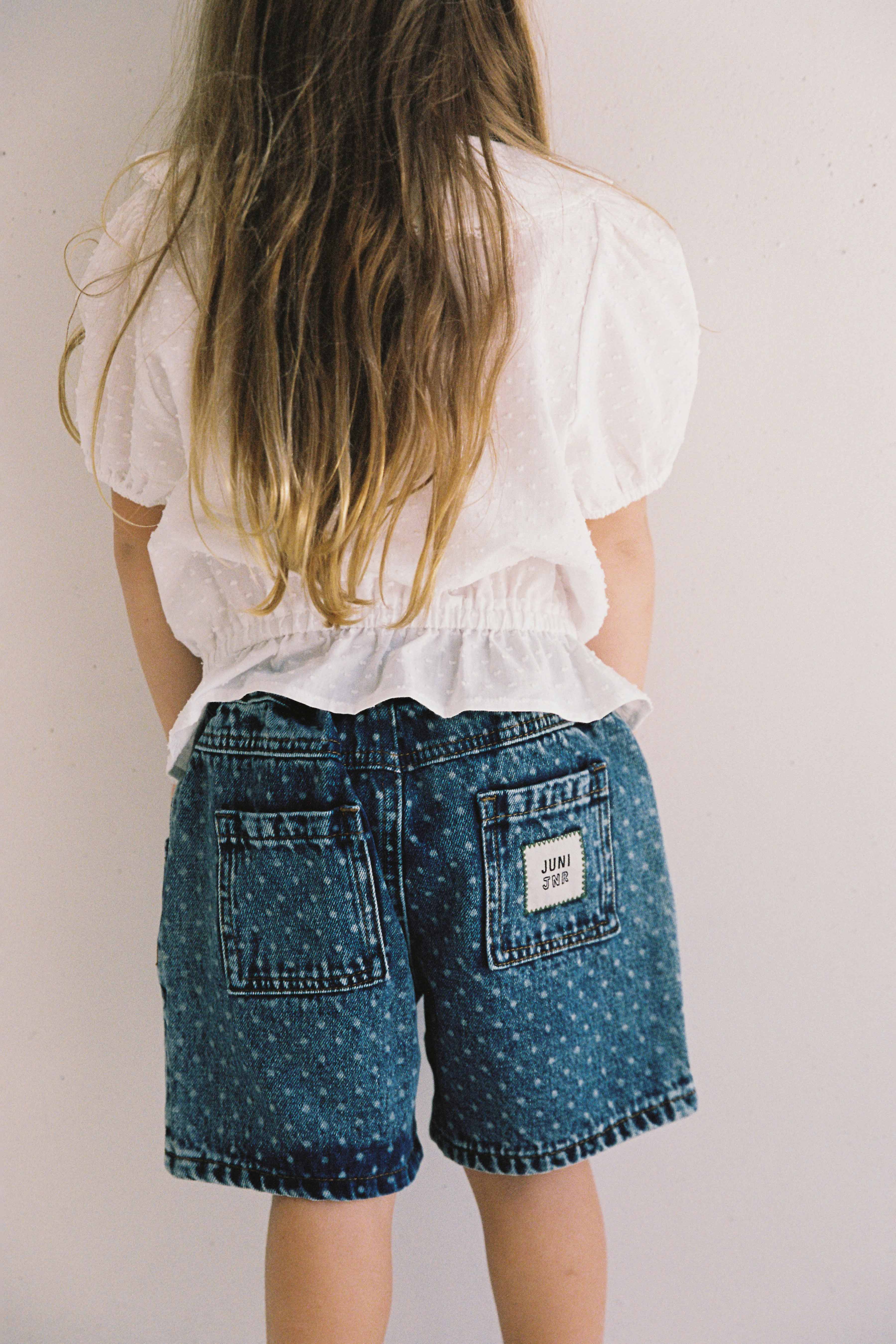 A young child with long, light brown hair stands facing a white wall. They are wearing a white, short-sleeved shirt and the Junior Bud Denim Short Spot by JUNI JUNIOR, featuring high-waisted 100% cotton denim with an elastic waistband and patchwork on the back pockets. The label on the right pocket reads "JUNI.