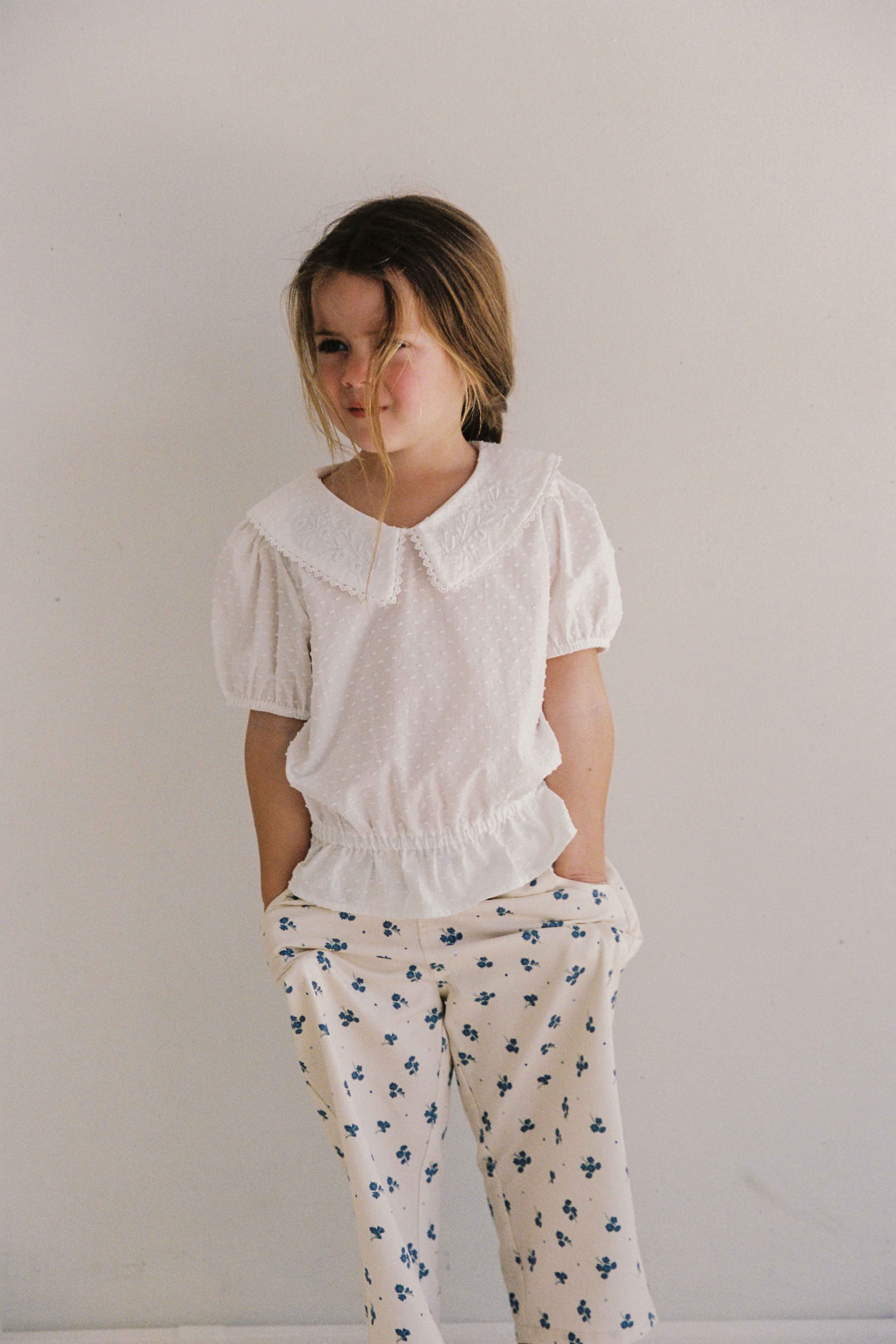 A young girl with blonde hair wearing the Junior Celine Blouse by JUNI JUNIOR, a white blouse featuring a Peter Pan collar and short puffed sleeves made from ethically sourced cotton dobby fabric, along with white pants patterned with blue flowers, stands against a plain white background with her hands in her pockets, looking off to the side.