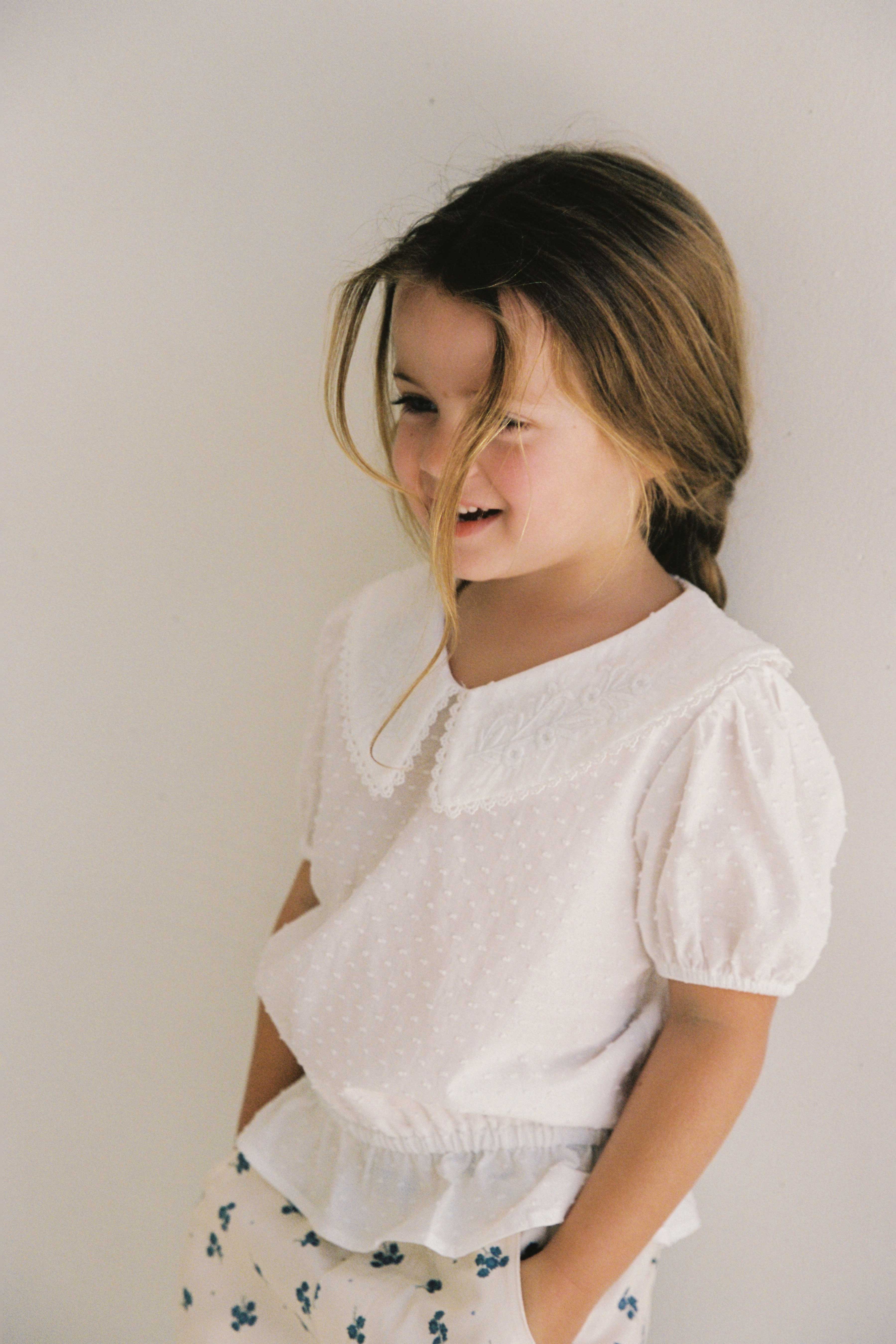 A young girl with long brown hair stands against a white background. She is wearing the Junior Celine Blouse from JUNI JUNIOR, which features a large collar and short, puffed sleeves made from soft cotton dobby. Paired with patterned pants, she has a soft smile on her face as a lock of hair falls over her forehead.