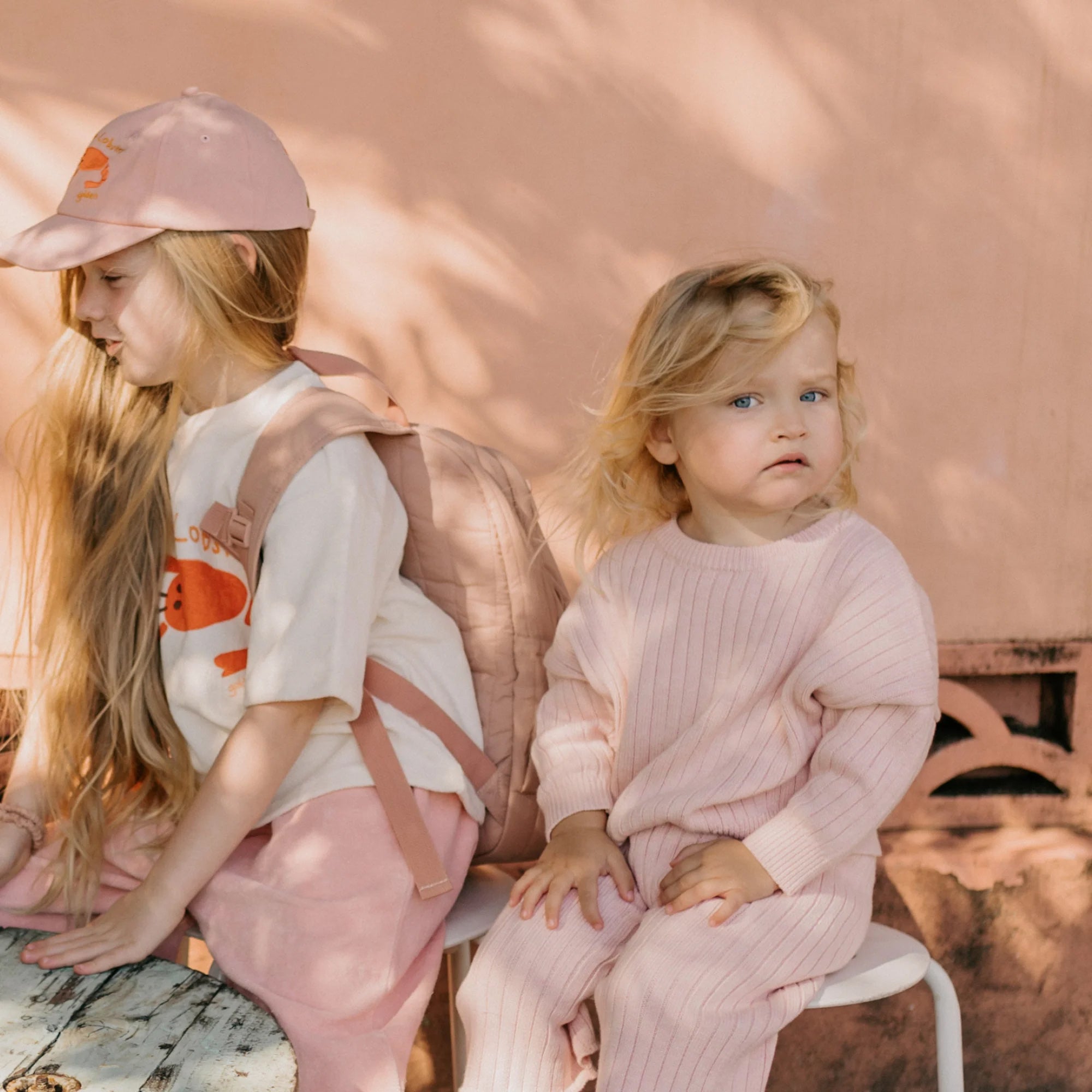 Two young children are sitting outdoors. The child on the left has long hair, wears a pink cap, a light-colored t-shirt with a graphic, pink pants, and a matching backpack. The child on the right, dressed in a relaxed fit GOLDEN CHILDREN Junee Knit Sweater Macaron made from 100% cotton knit, sits on a white chair against a peach wall.