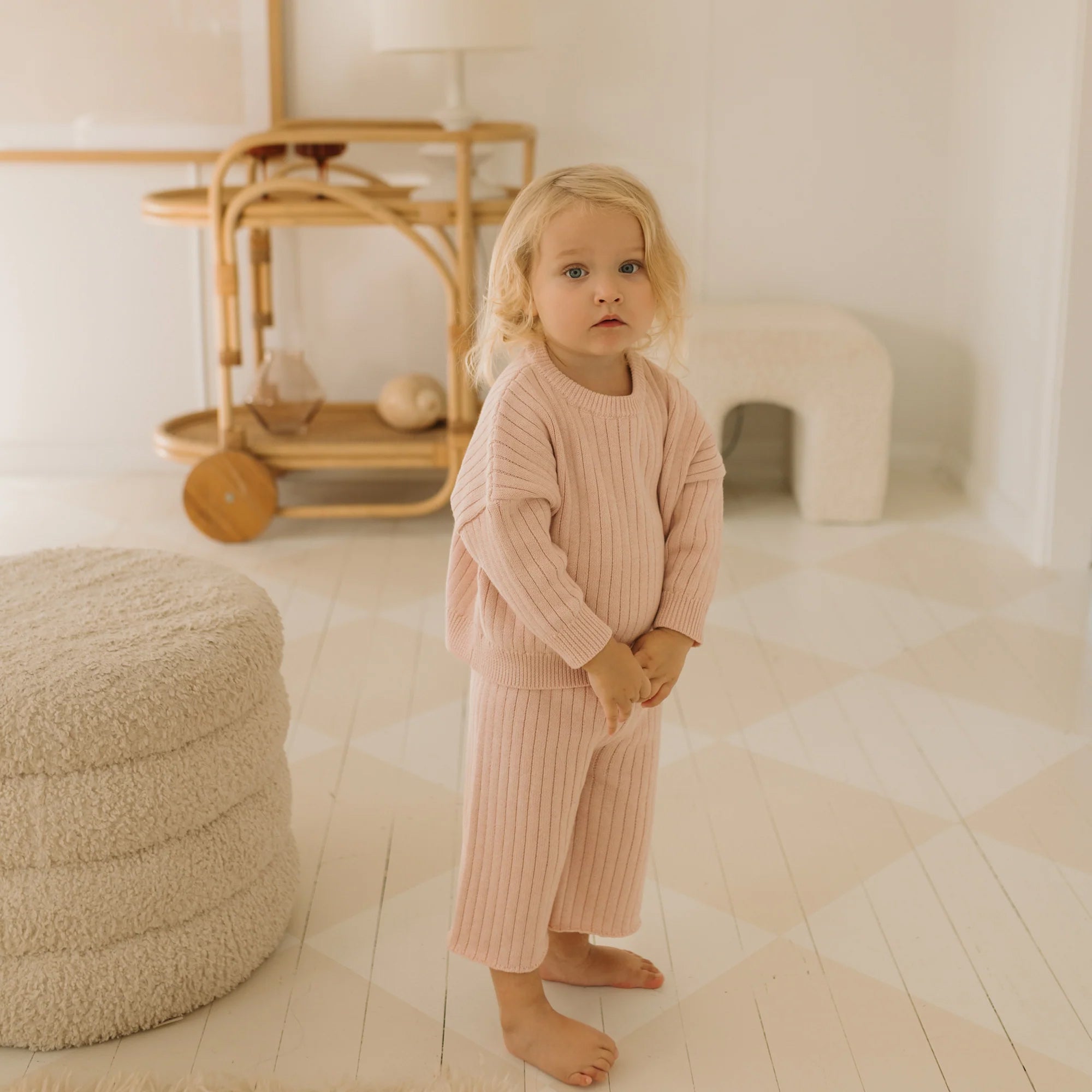 A young child with long blonde hair stands barefoot in a minimalistic room adorned with light-colored decor. The child is wearing a matching Junee Knit Sweater Macaron set made from 100% cotton knit and offering a relaxed fit by GOLDEN CHILDREN. Beside the child is a textured, round ottoman, and in the background, there's a wooden bar cart.