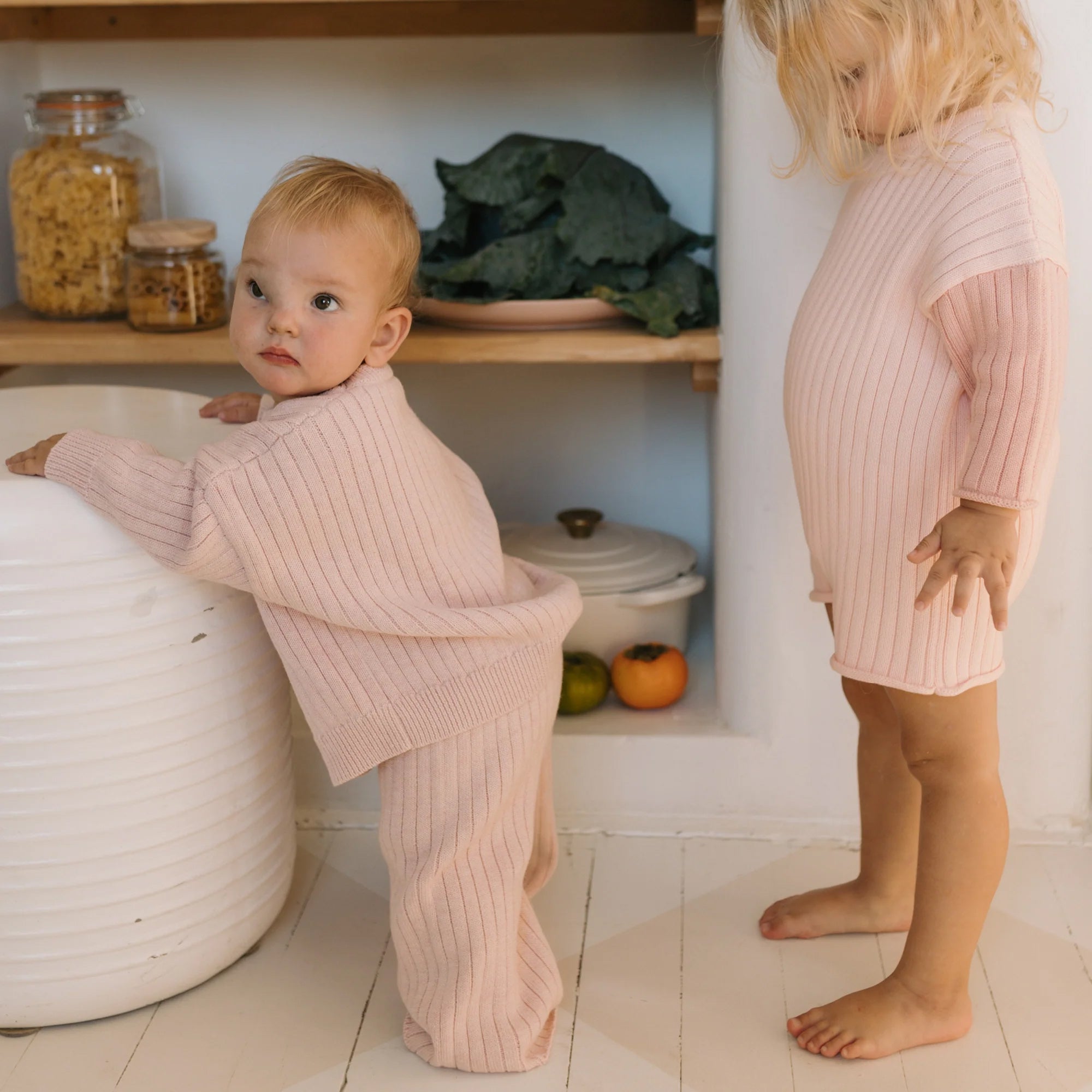 Two toddlers in ribbed, pastel-colored Junee Knit Sweaters Macaron by GOLDEN CHILDREN are standing indoors by pantry shelves. One child, with light hair, leans on a jar, while the other, with slightly longer light hair, stands beside some fresh produce and kitchen items on a white wooden floor.