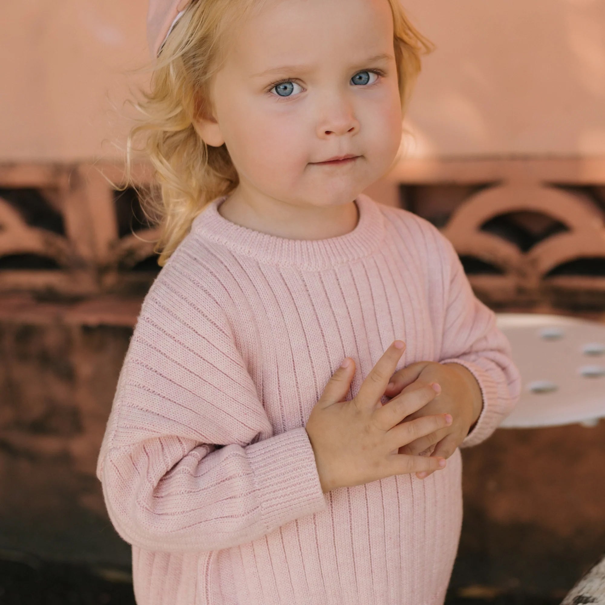 A young child with blonde hair and blue eyes, wearing a light pink Junee Knit Sweater Macaron from GOLDEN CHILDREN, stands outdoors. The 100% cotton knit sweater has a relaxed fit and drapes comfortably as the child clasps their hands together and looks directly at the camera. A soft-focus, peach-colored wall and part of a white patterned bench complete the background.