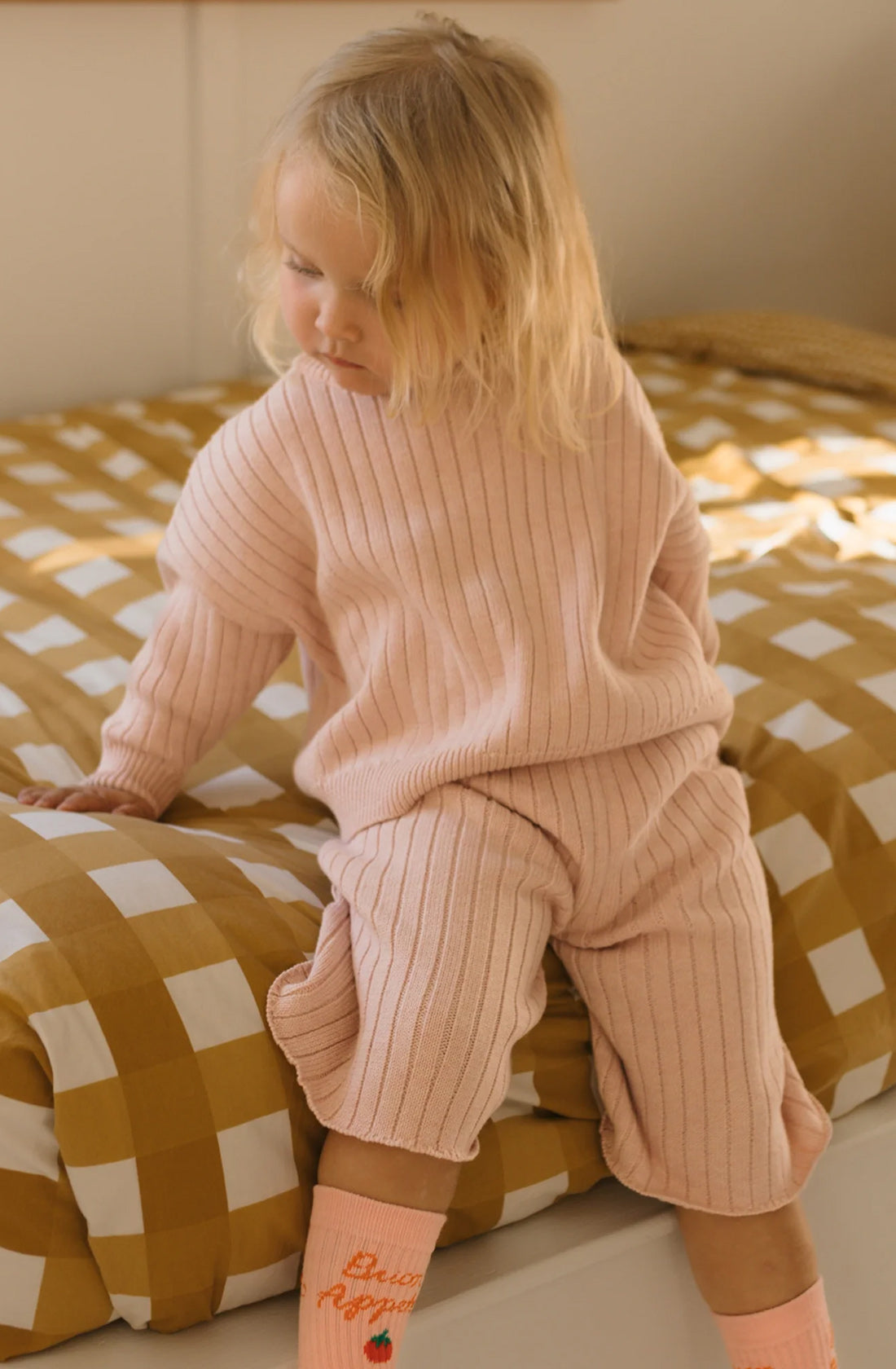 A young child with blonde hair sits on the edge of a bed, wearing a relaxed fit Junee Knit Sweater Macaron from GOLDEN CHILDREN and matching pants, both crafted from 100% cotton knit. The bed has a brown and white checkered blanket, and the child is looking down, with one leg dangling off the bed.