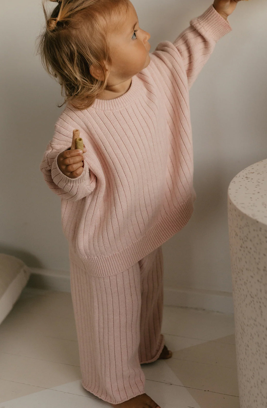 A toddler stands indoors wearing a cozy, oversized pink Junee Knit Sweater Macaron and matching pants from GOLDEN CHILDREN. They have light-colored hair and are reaching up towards something out of the frame with one hand while holding a small item in the other. The 100% cotton knit provides a soft, relaxed fit perfect for little explorers.