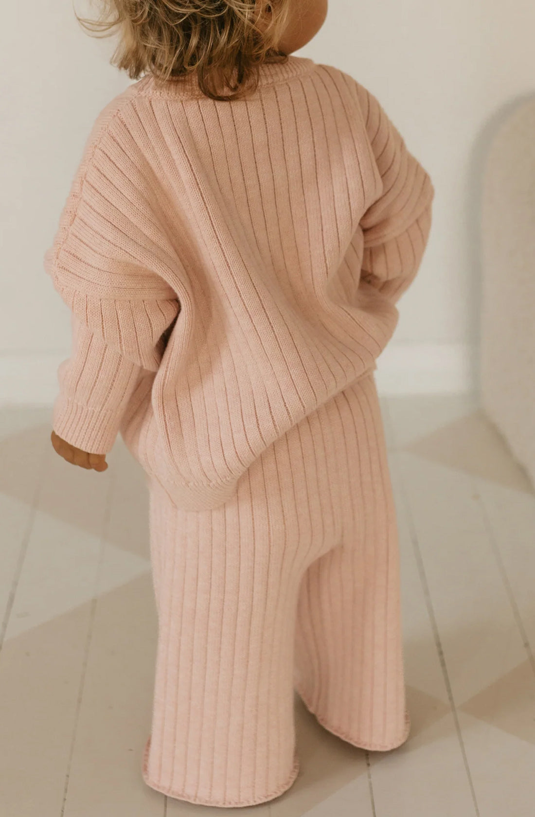 A child with curly hair is standing and facing away from the camera. They are wearing a cozy, ribbed, peach-colored Junee Knit Sweater Macaron by GOLDEN CHILDREN and matching pants. The relaxed fit complements the soft, neutral-colored background and light flooring of the room.